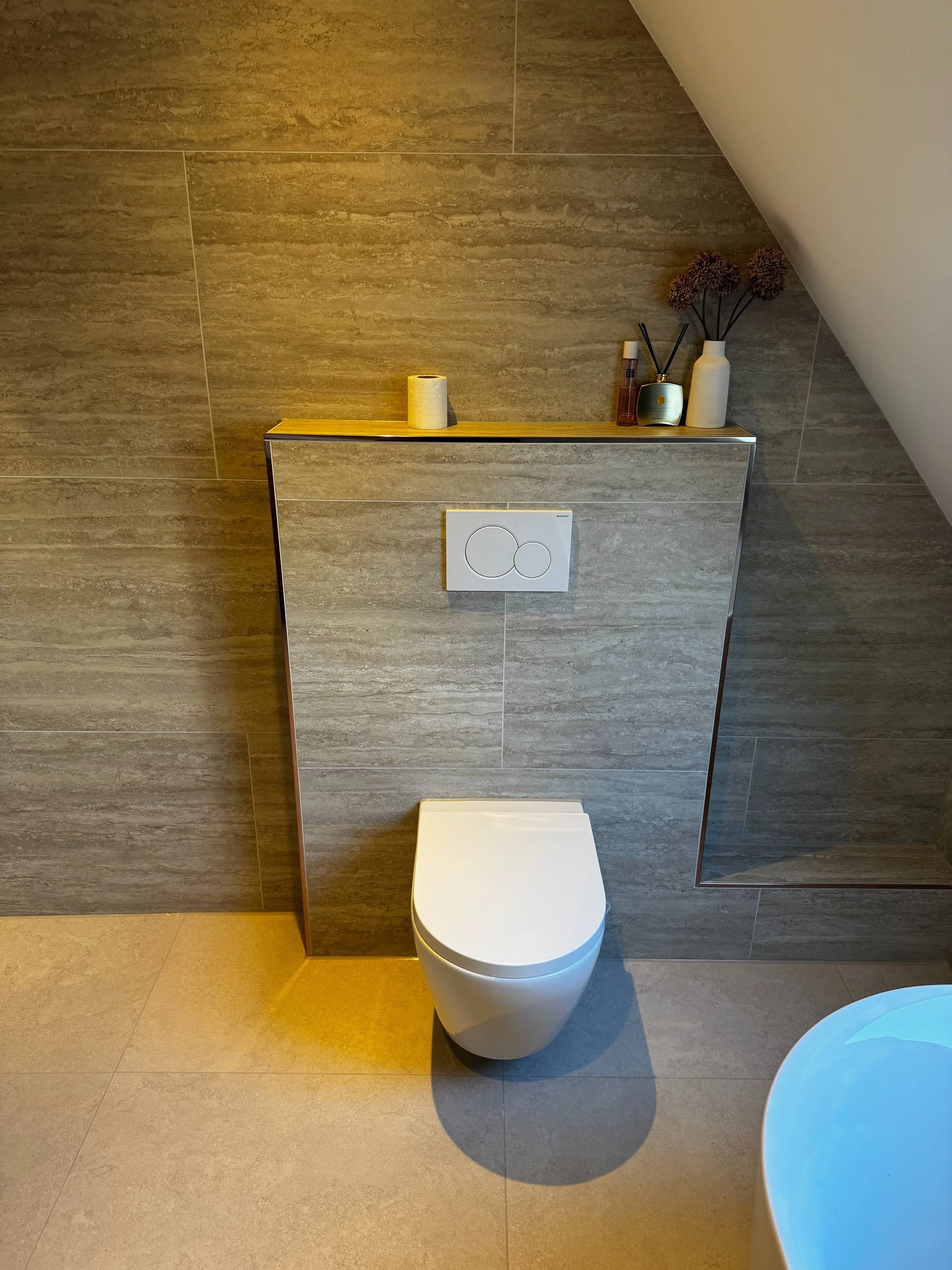 Modern toilet in een badkamer met grote grijze tegels, decoratieve vazen en geurstokjes op een plank boven de toilet.