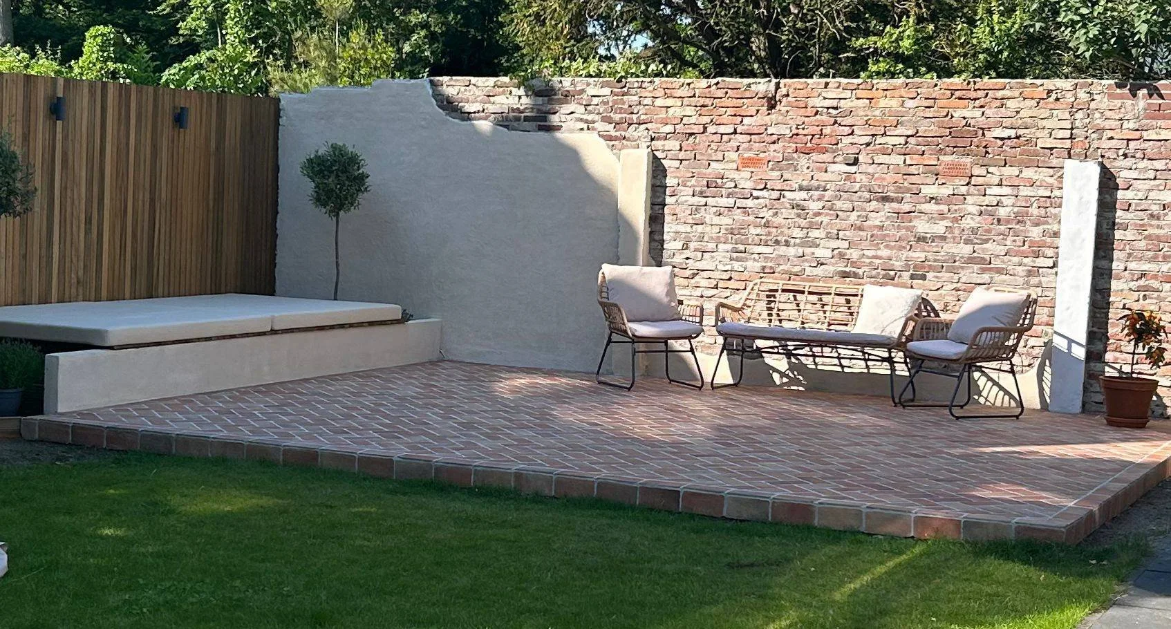 Een rustige tuin met een betegeld terras, een bank met kussens, twee stoelen met kussens, een muur van bakstenen, een houten hek en groene planten.