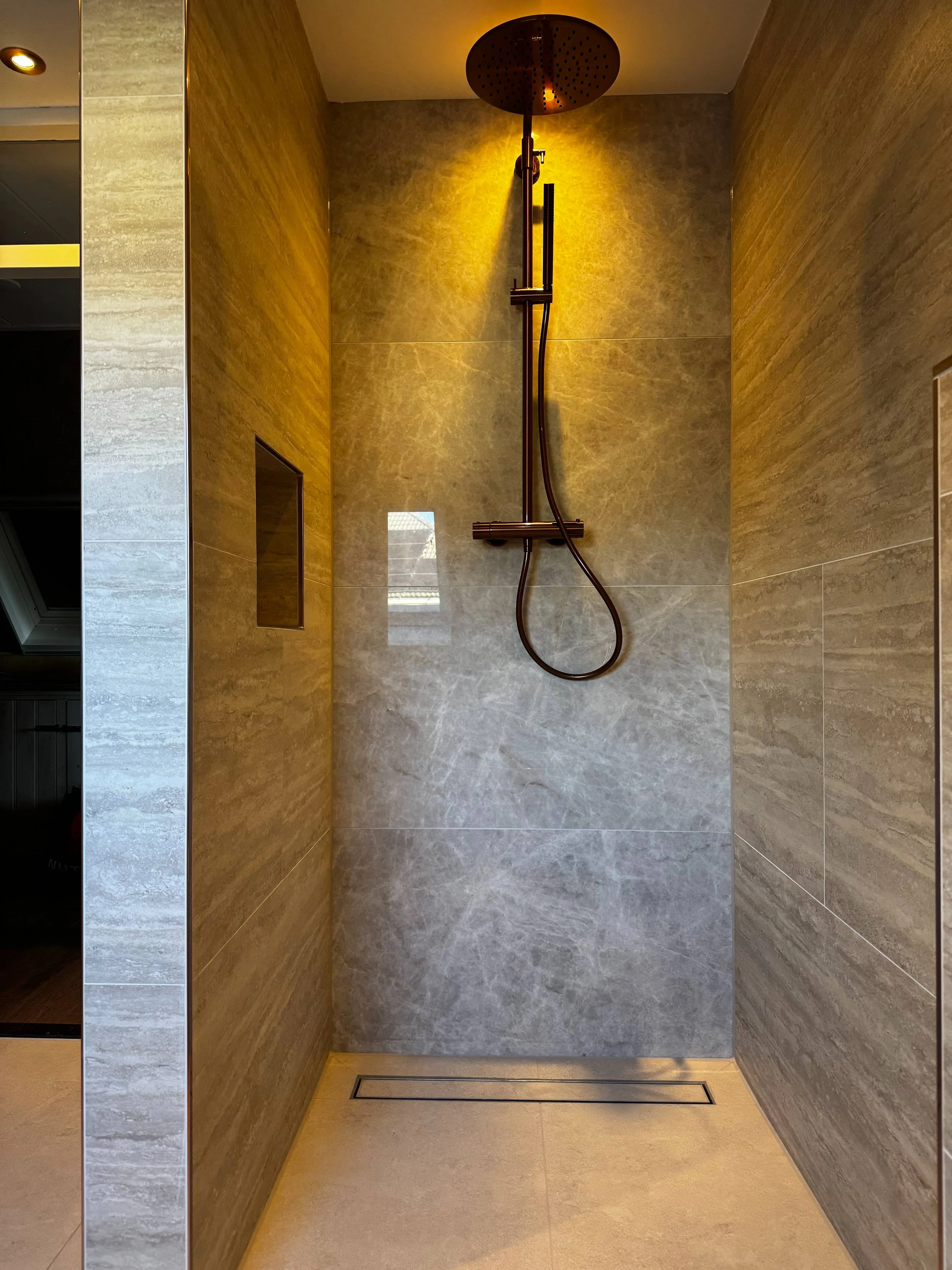 Moderne inloopdouche met een hoofddouche en wandbediening, betegeld met grote, grijze natuurstenen tegels, met een kleine raam en inbouwafvoer in de vloer.