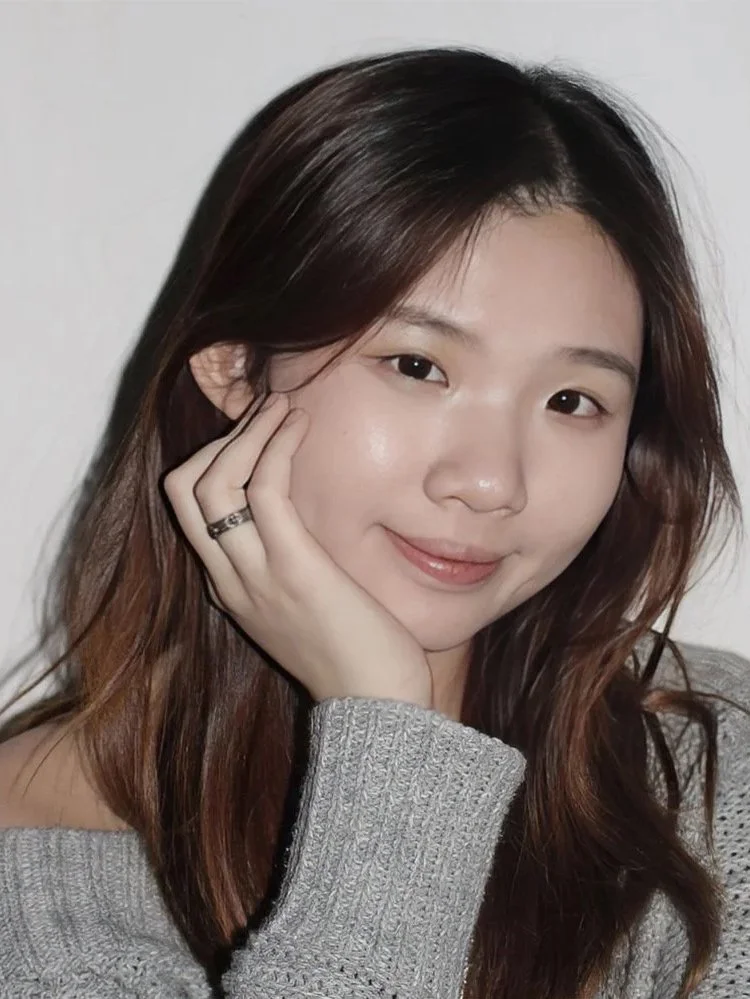 A young woman with medium-length brown hair and fair skin, smiling gently, resting her head on her hand, wearing a gray knit sweater.