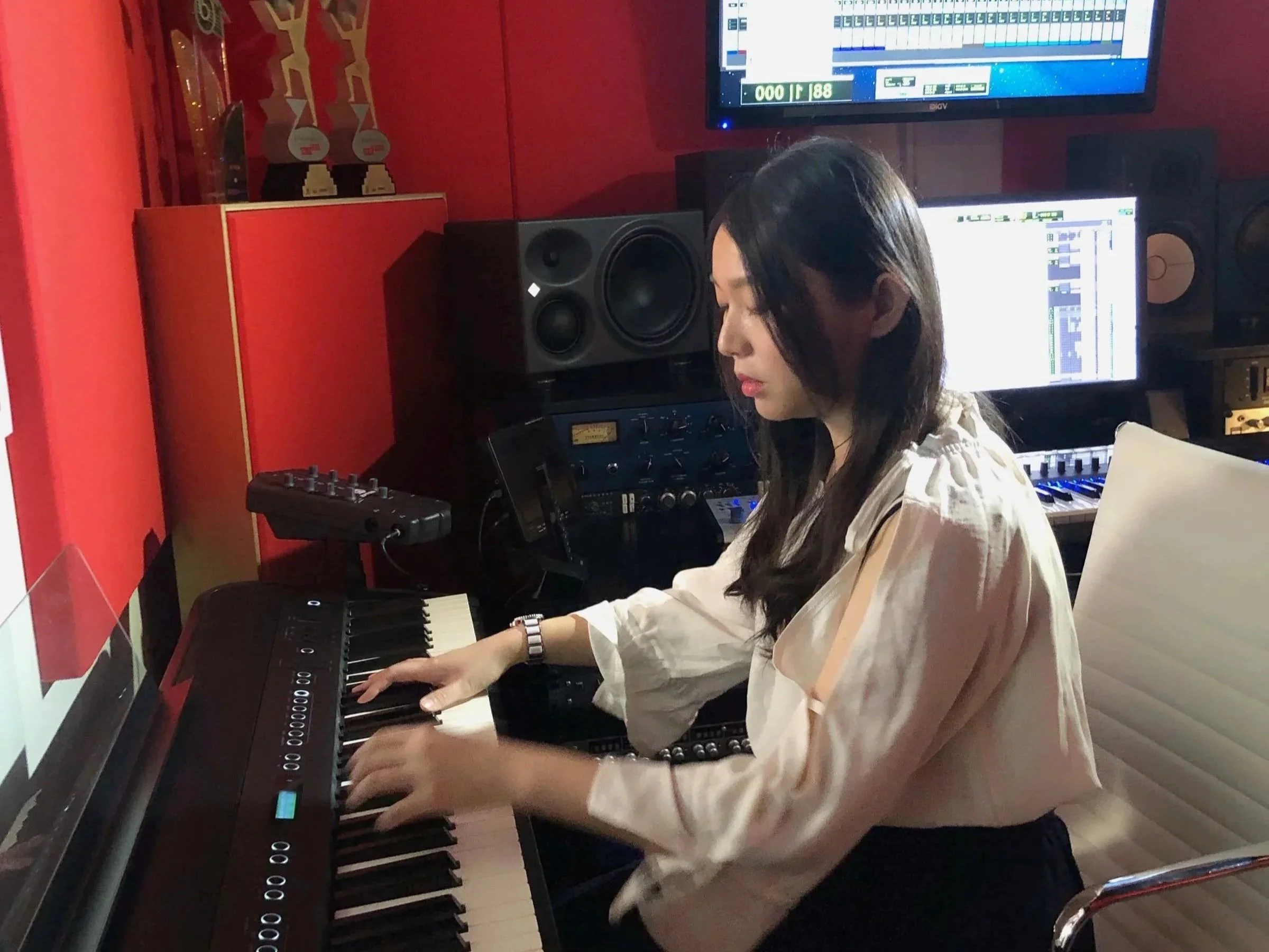A woman with long dark hair and a white blouse playing a digital keyboard in a music studio. There are two large computer monitors displaying music production software, and black studio speakers on a red wall behind her. Trophies are visible on a shelf above the monitors.