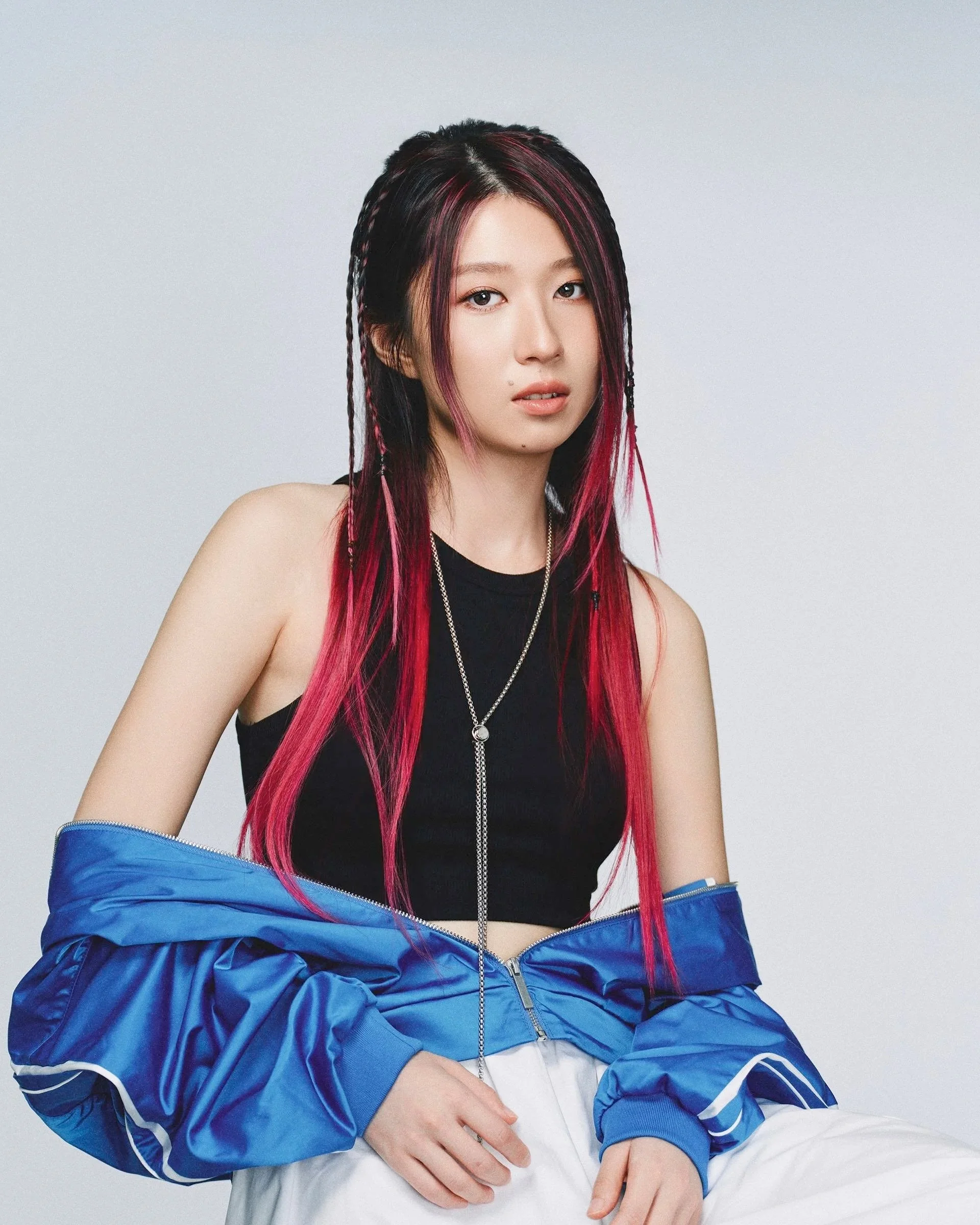 Young woman with pink and black braided hair, wearing a black tank top, layered necklaces, a blue jacket partially unzipped, and white pants, standing against a plain gray background.