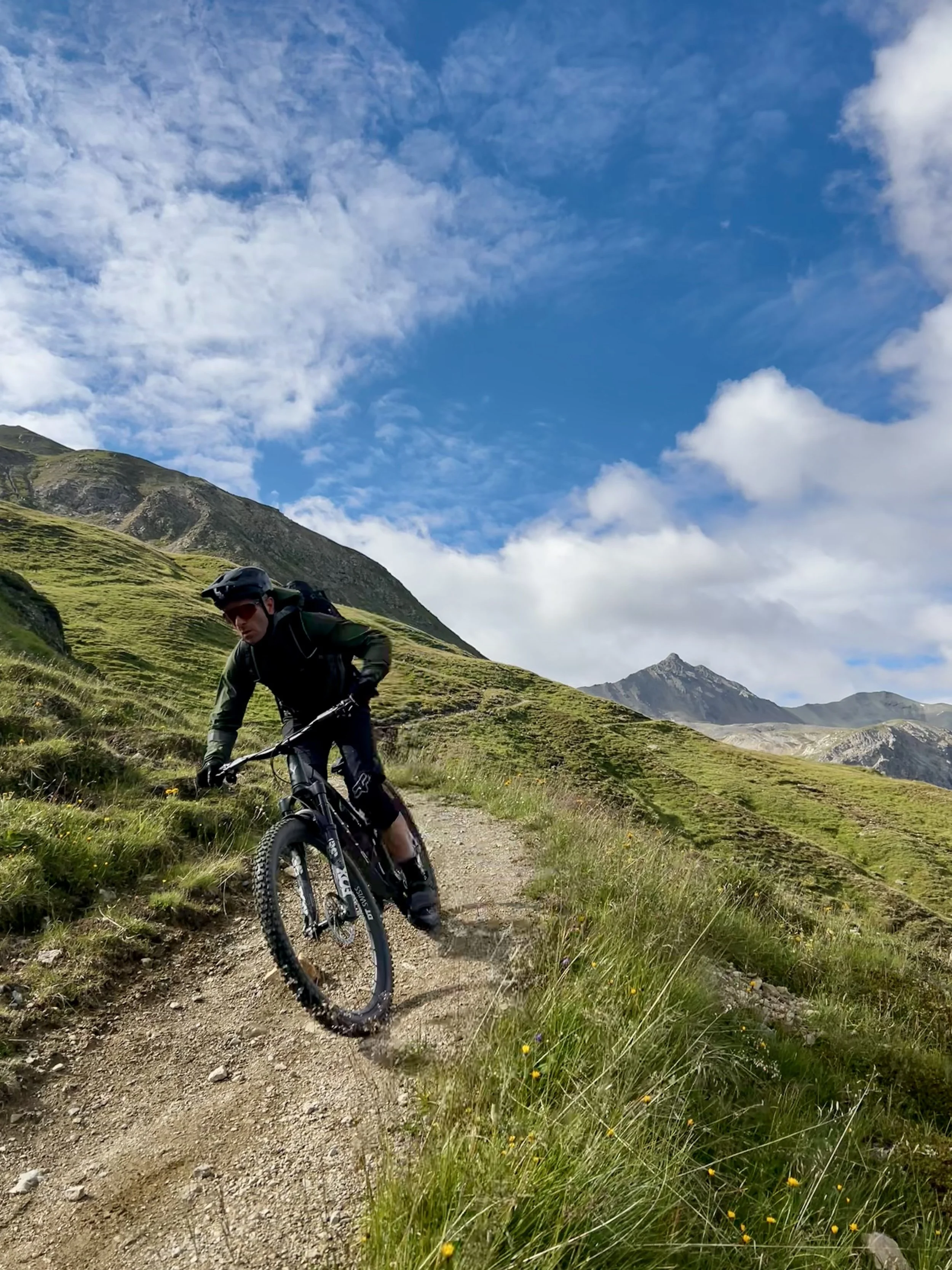 Ein Mountainbiker fährt auf einem schmalen Wanderweg in einer grünen, bergigen Landschaft unter einem blauen Himmel mit Wolken.