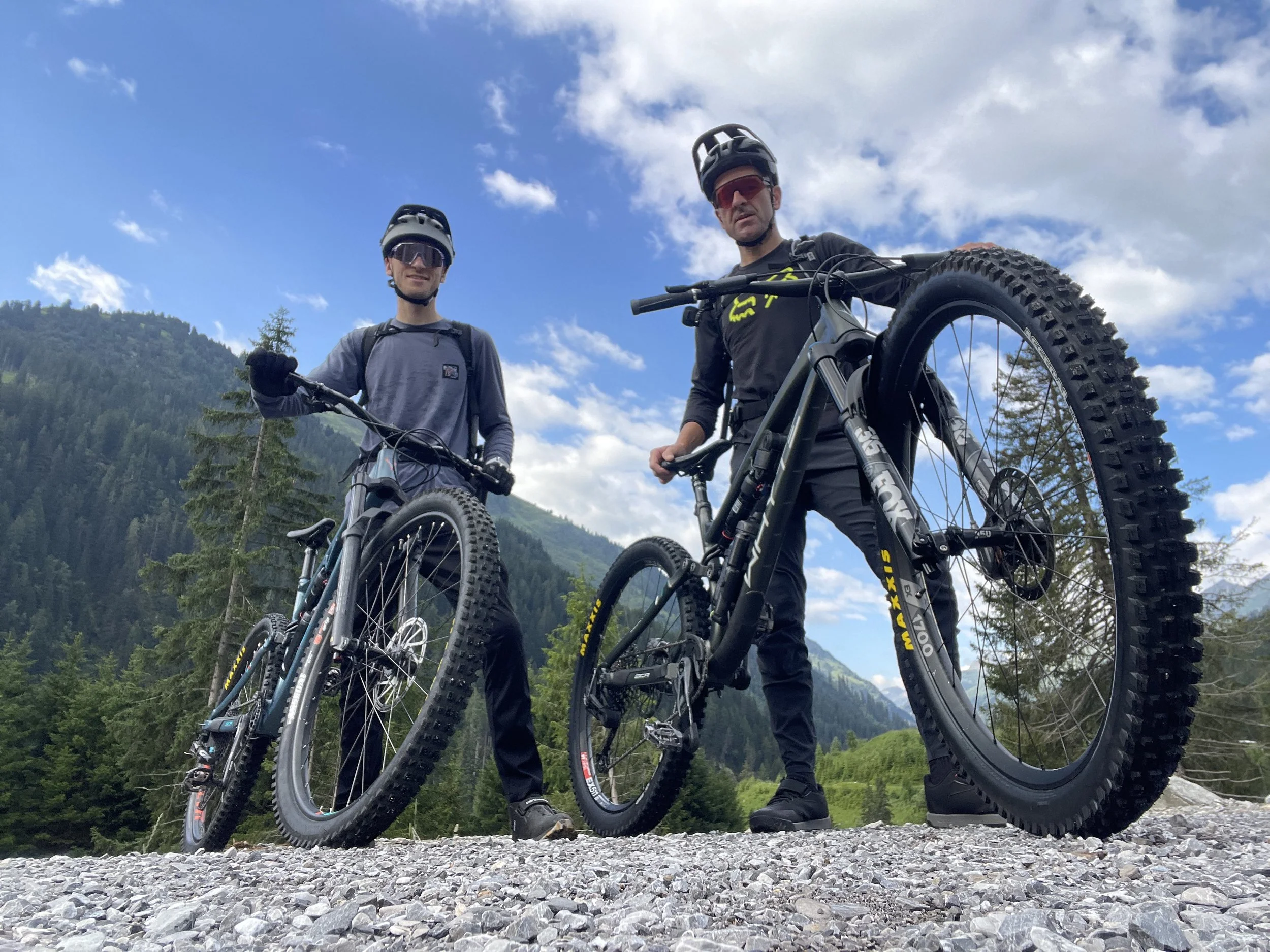 Zwei Mountainbiker stehen mit ihren Fahrrädern auf einer Felsstrecke vor einem grünen Gebirge unter einem blauen Himmel mit weißen Wolken.