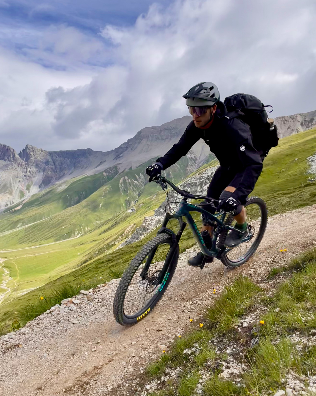 Ein Mountainbiker fährt auf einer schmalen, felsigen Bergstraße in einer alpinen Landschaft mit grünen Tälern und hohen Bergen im Hintergrund.