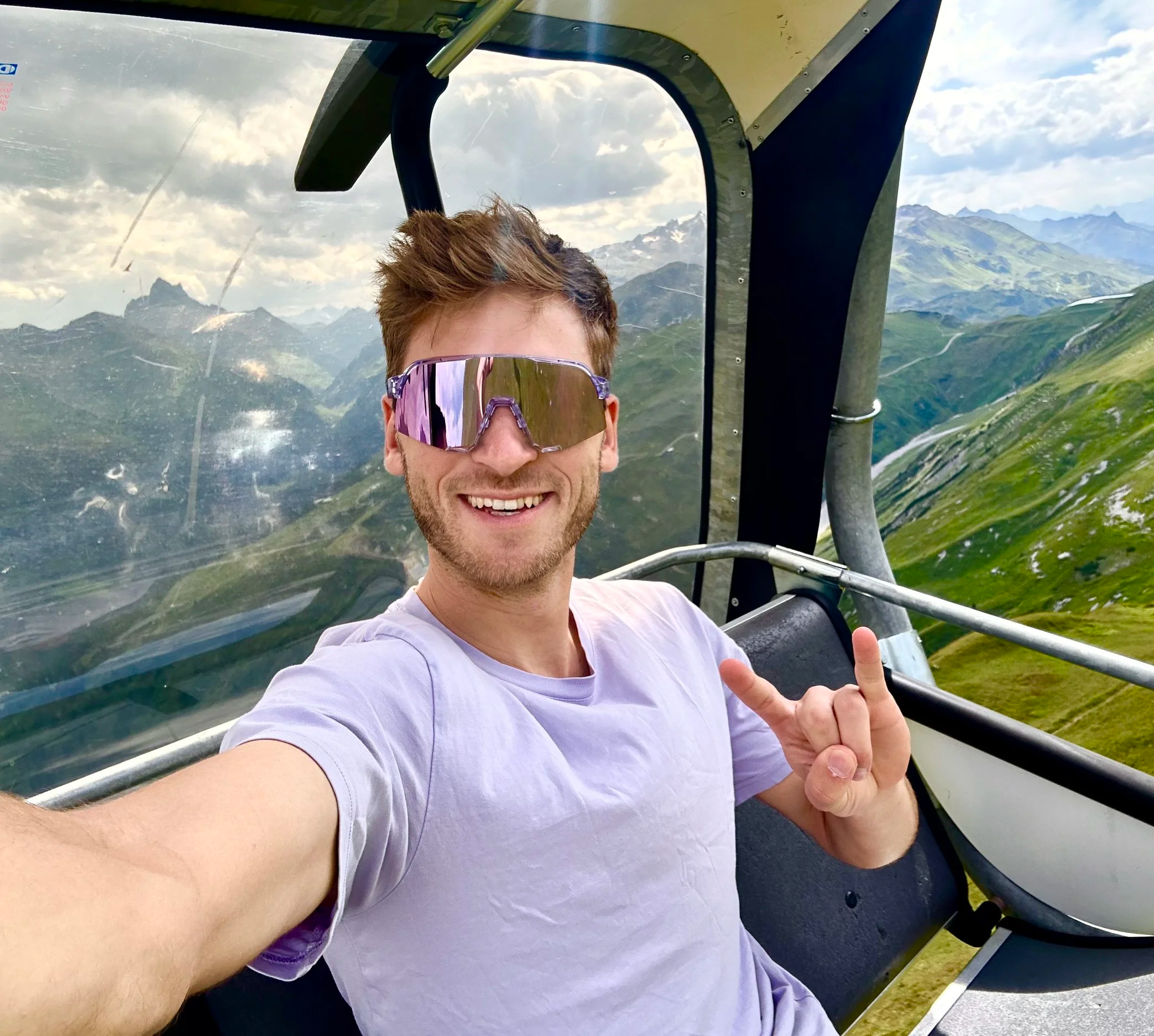 Junger Mann in einer Gondel mit Berglandschaft im Hintergrund, trägt Sonnenbrille und macht ein Peace-Zeichen.