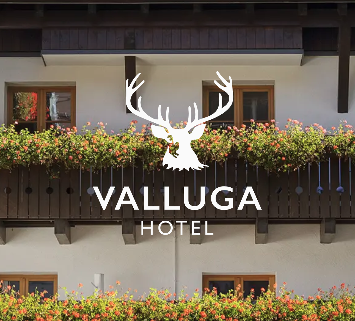 Bild eines Hotels mit Balkon und Blumenkästen, das Logo des Valluga Hotels mit einem Hirschkopf und Geweih ist darüber sichtbar.