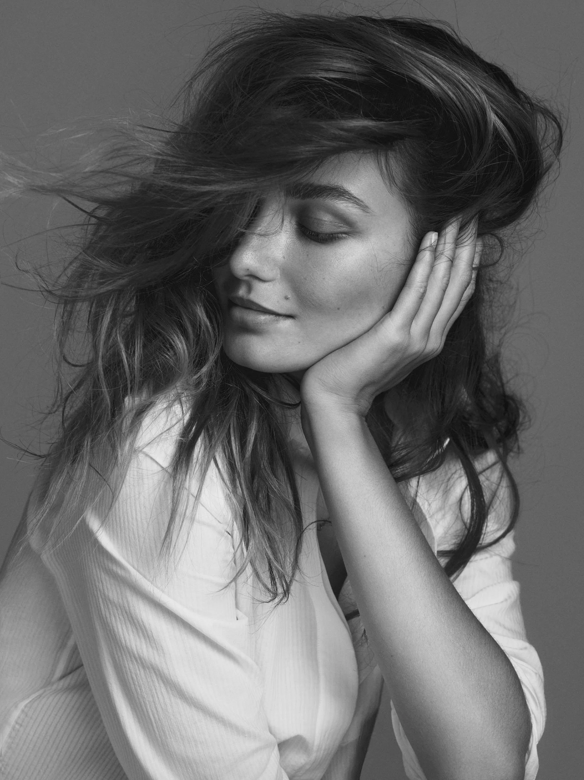 Black and white portrait of a woman with long, wavy hair, eyes closed, gently resting her face on her hand, wearing a light-colored shirt.