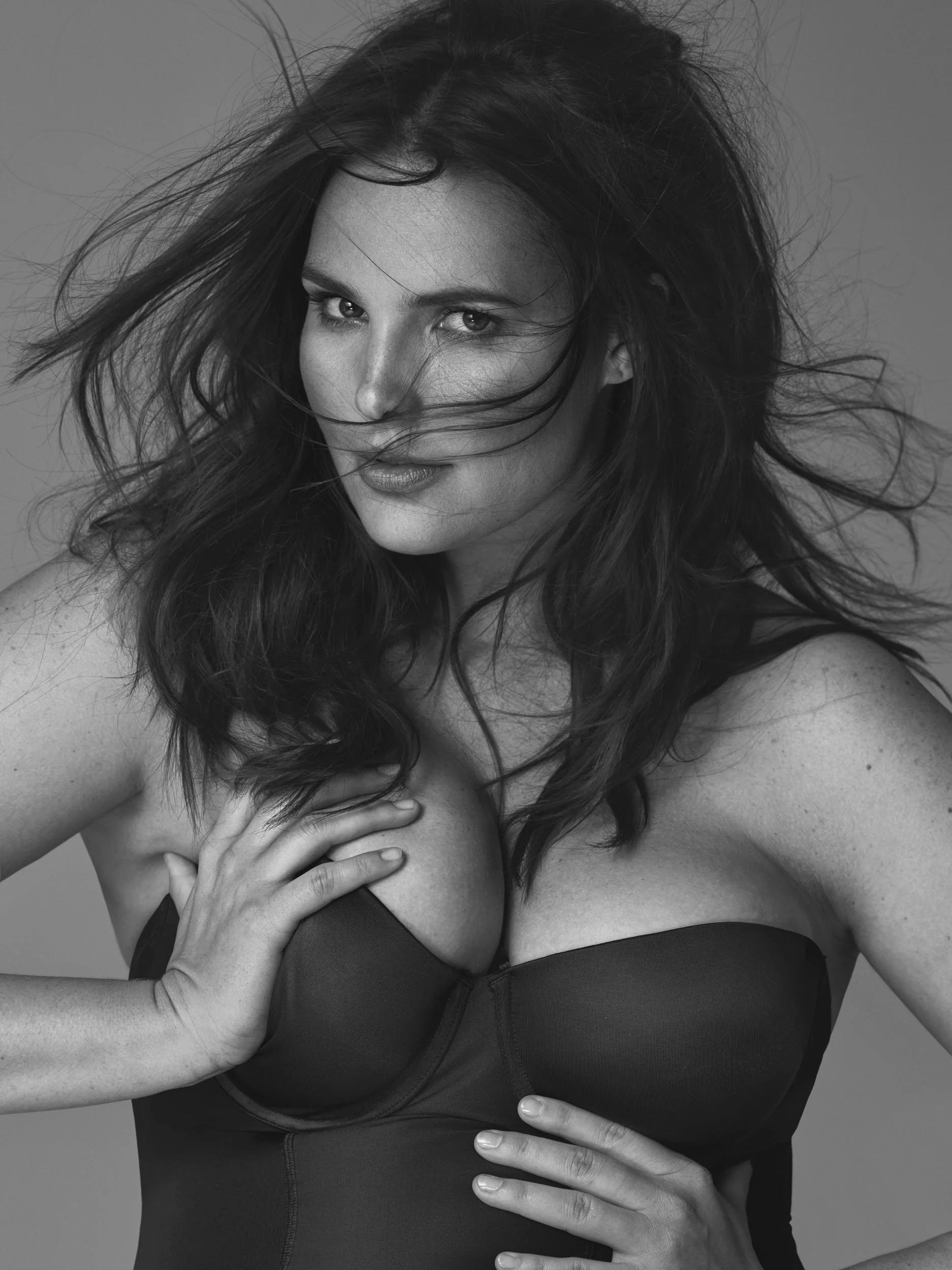 Black and white photo of a woman with flowing hair, wearing a strapless top. She is looking at the camera with a confident expression, her hand on her shoulder and her other hand resting on her waist.