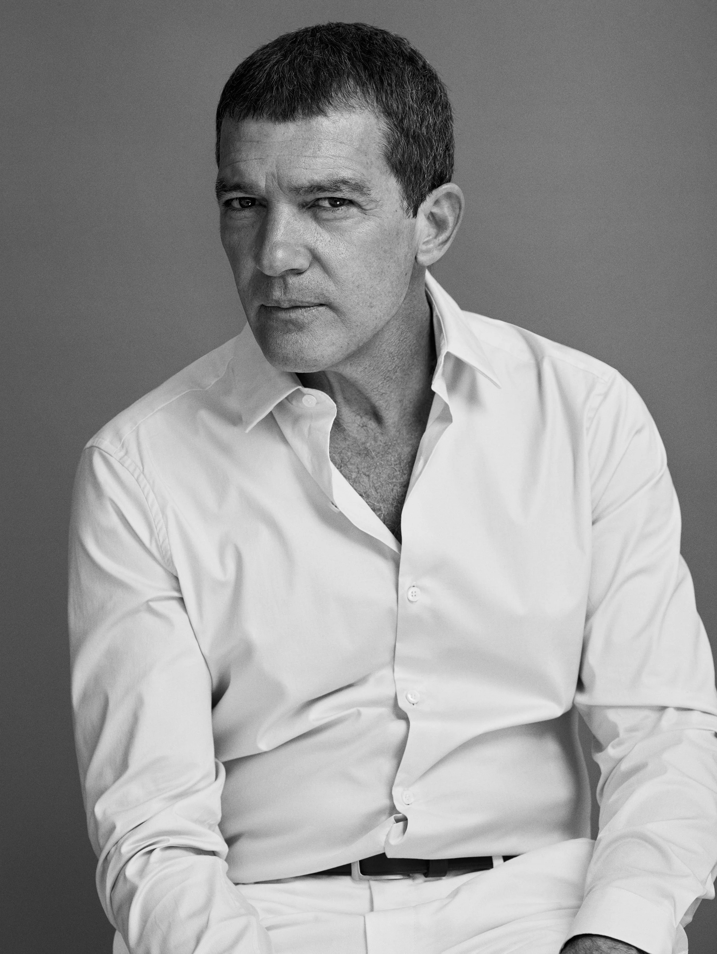 A black-and-white portrait of a man with short dark hair, wearing a white button-up shirt, looking directly at the camera with a serious expression.