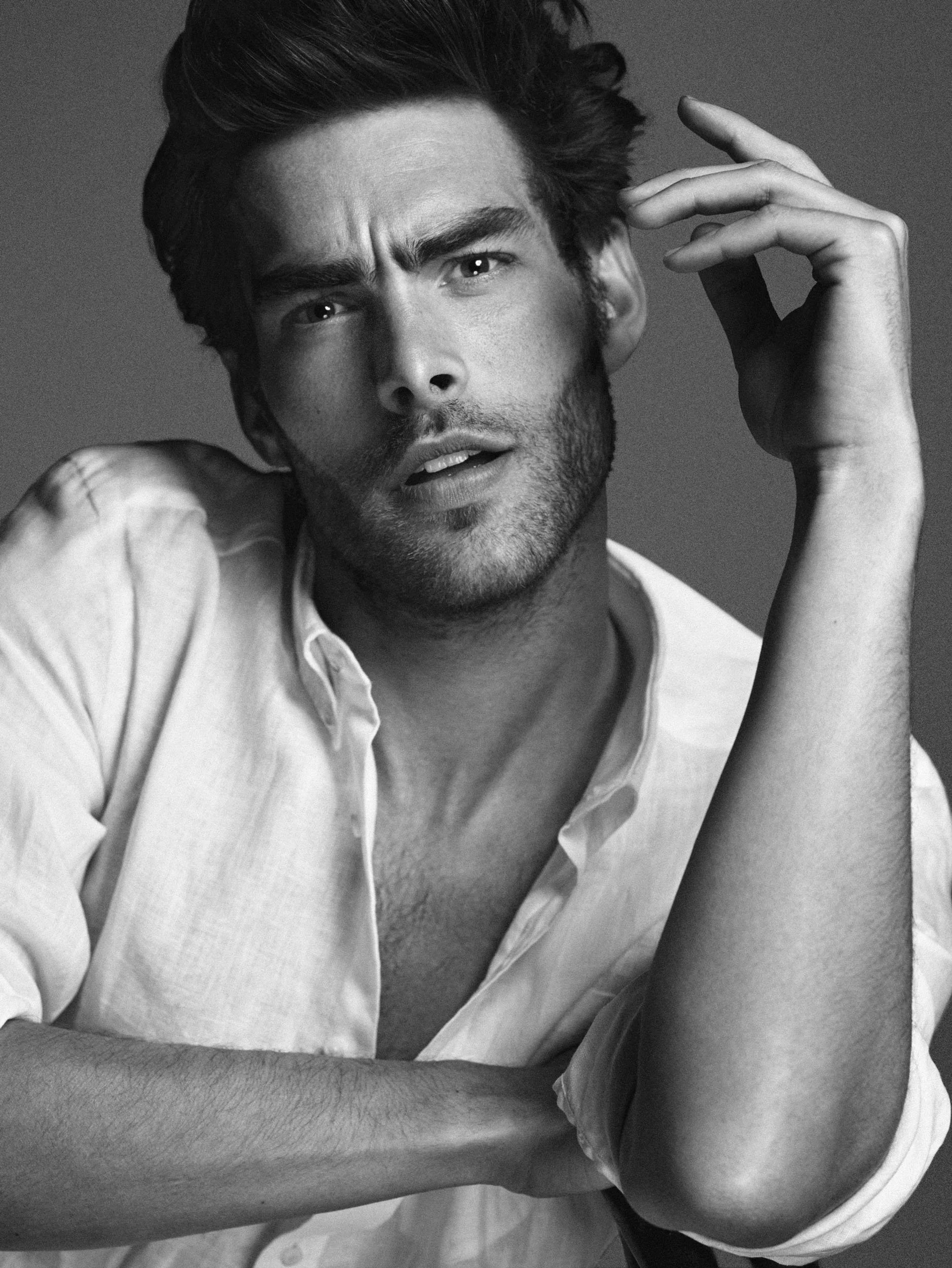 A black-and-white portrait of a man with dark, messy hair, a beard, and wearing a slightly unbuttoned white shirt. He has a serious expression, with one hand touching his hair and the other arm crossed over his chest.