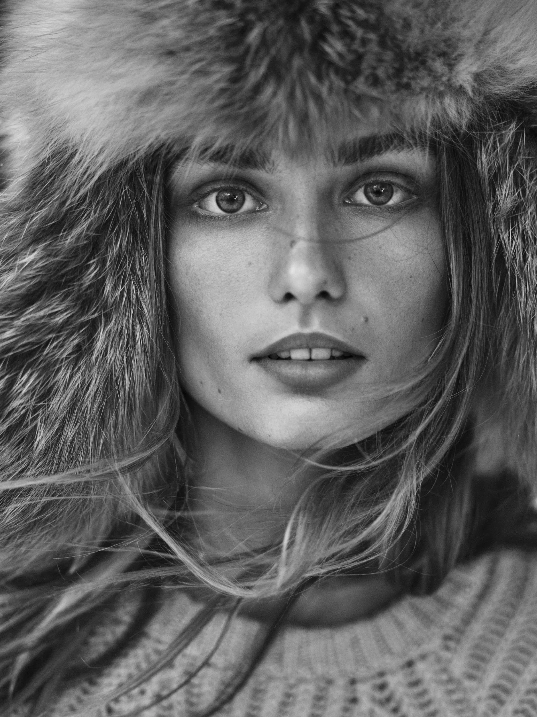 Close-up black and white photo of a woman with light eyes wearing a fur-lined hood and a knitted sweater.