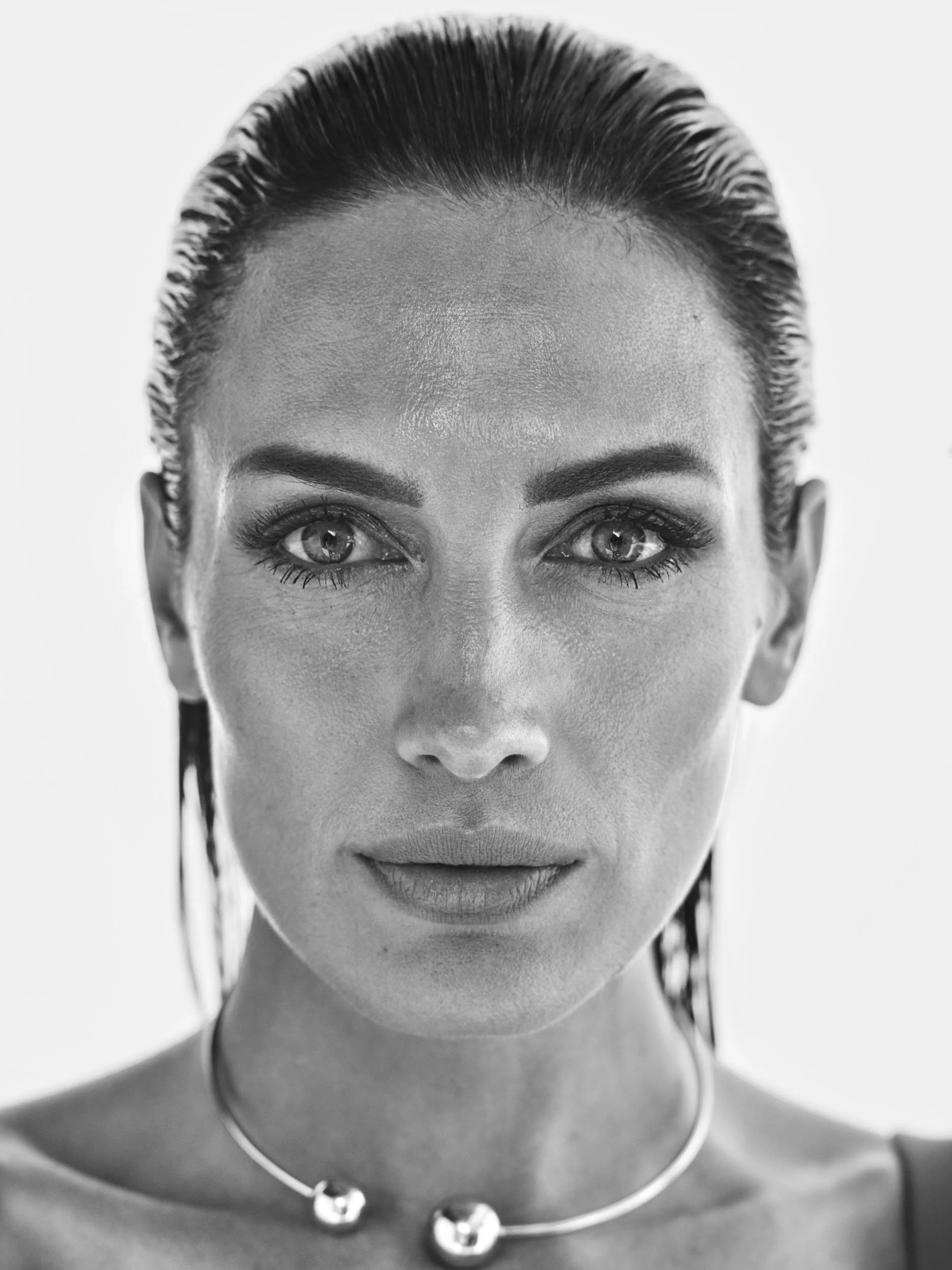 Close-up black and white portrait of a woman with wet hair, intense gaze, and wearing a necklace with two spherical pendants.
