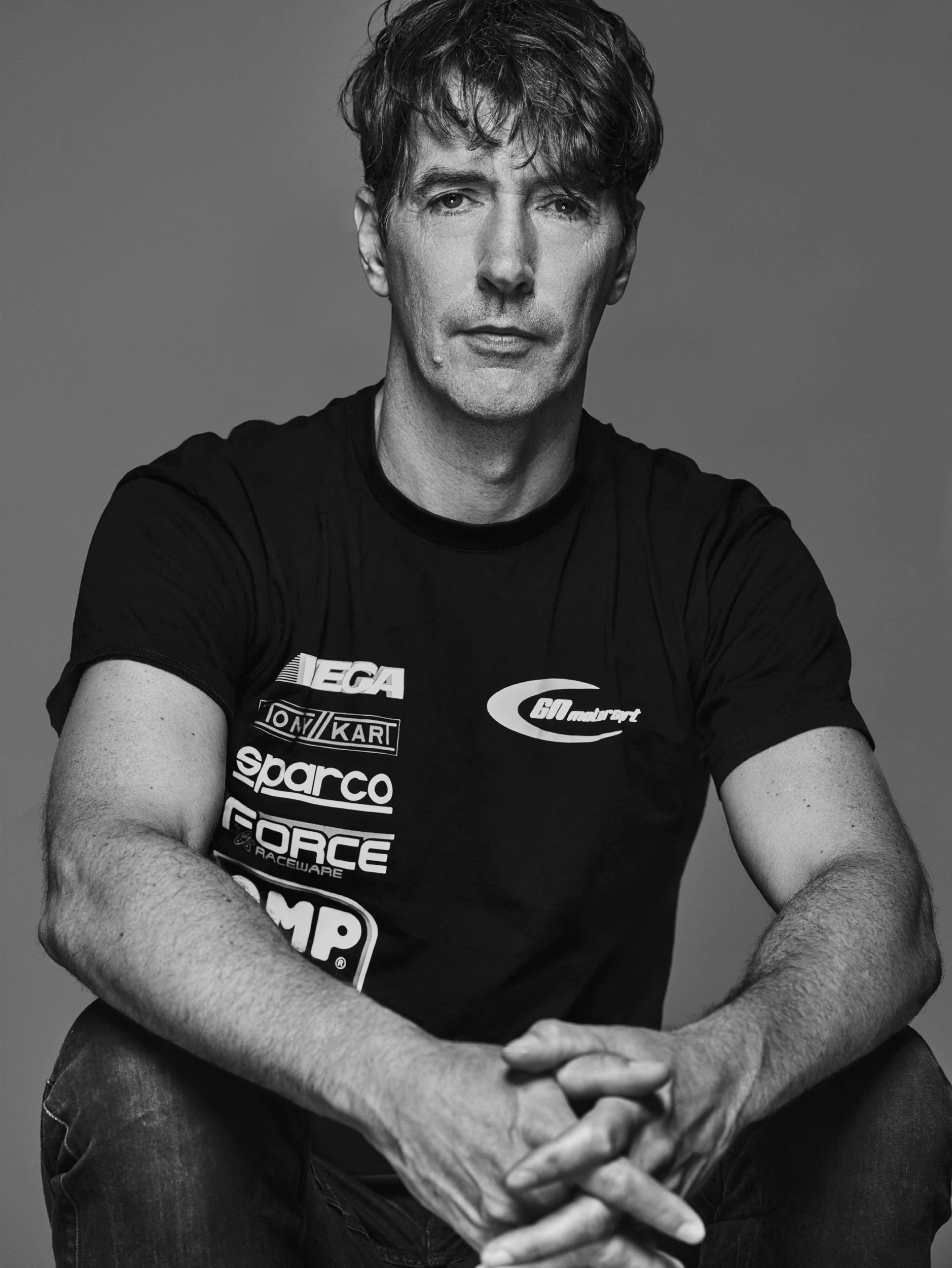 Black and white portrait of a man with messy hair, sitting with his hands clasped in front of him, wearing a T-shirt with racing logos such as EA, Tony Kart, Sparco, and Force.