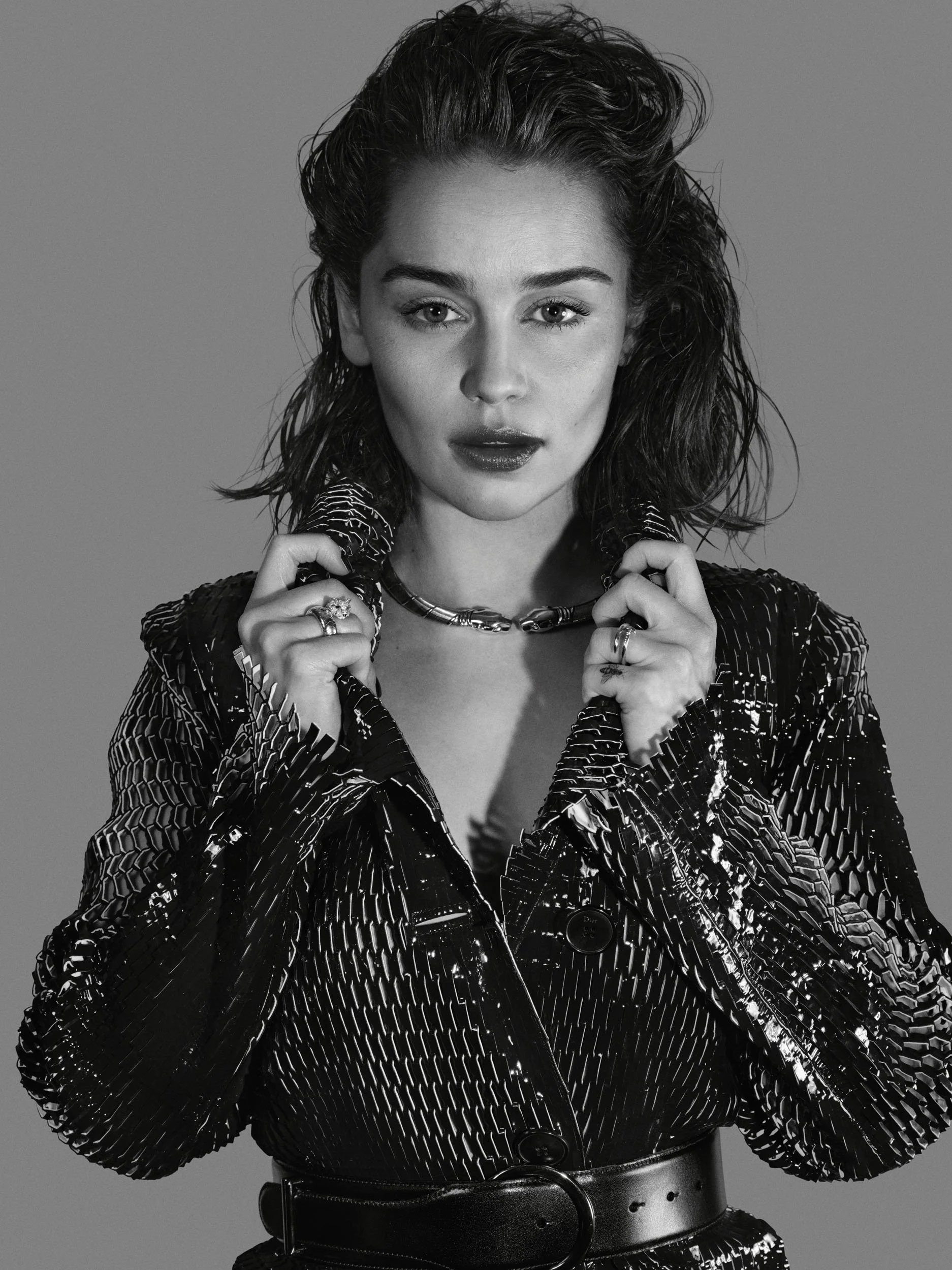 Black and white photo of woman with dark, wavy hair wearing a shiny, textured jacket, holding a necklace, with a serious expression.