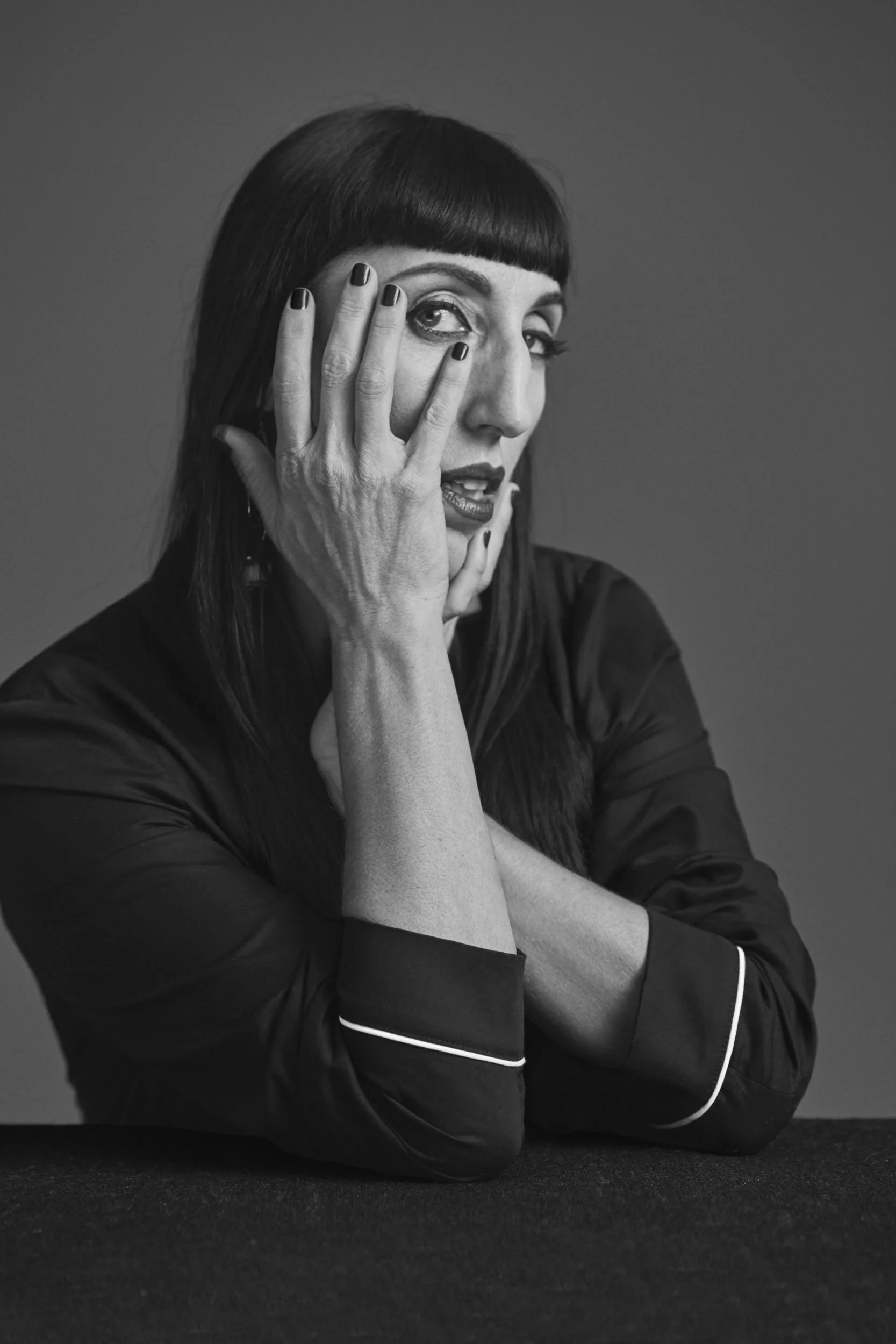 A woman with dark hair and bangs, wearing a silky black shirt with white piping, is sitting with her arms crossed on a dark surface. She has her left hand covering part of her face and her right hand resting on her cheek, looking thoughtfully to the 