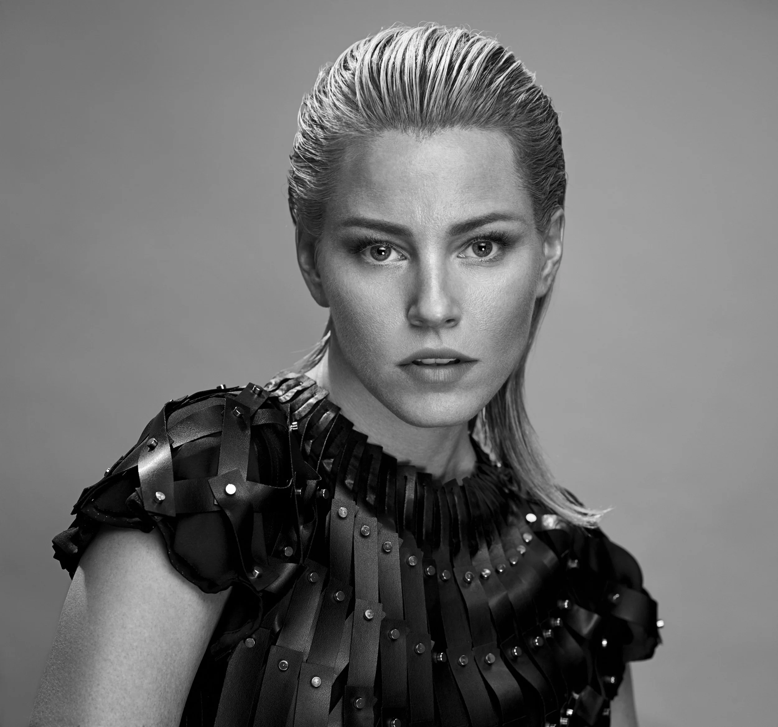 Black and white portrait of a woman with short, slicked-back hair, wearing a textured, shiny black top with metallic embellishments and ruffled details on the sleeves.