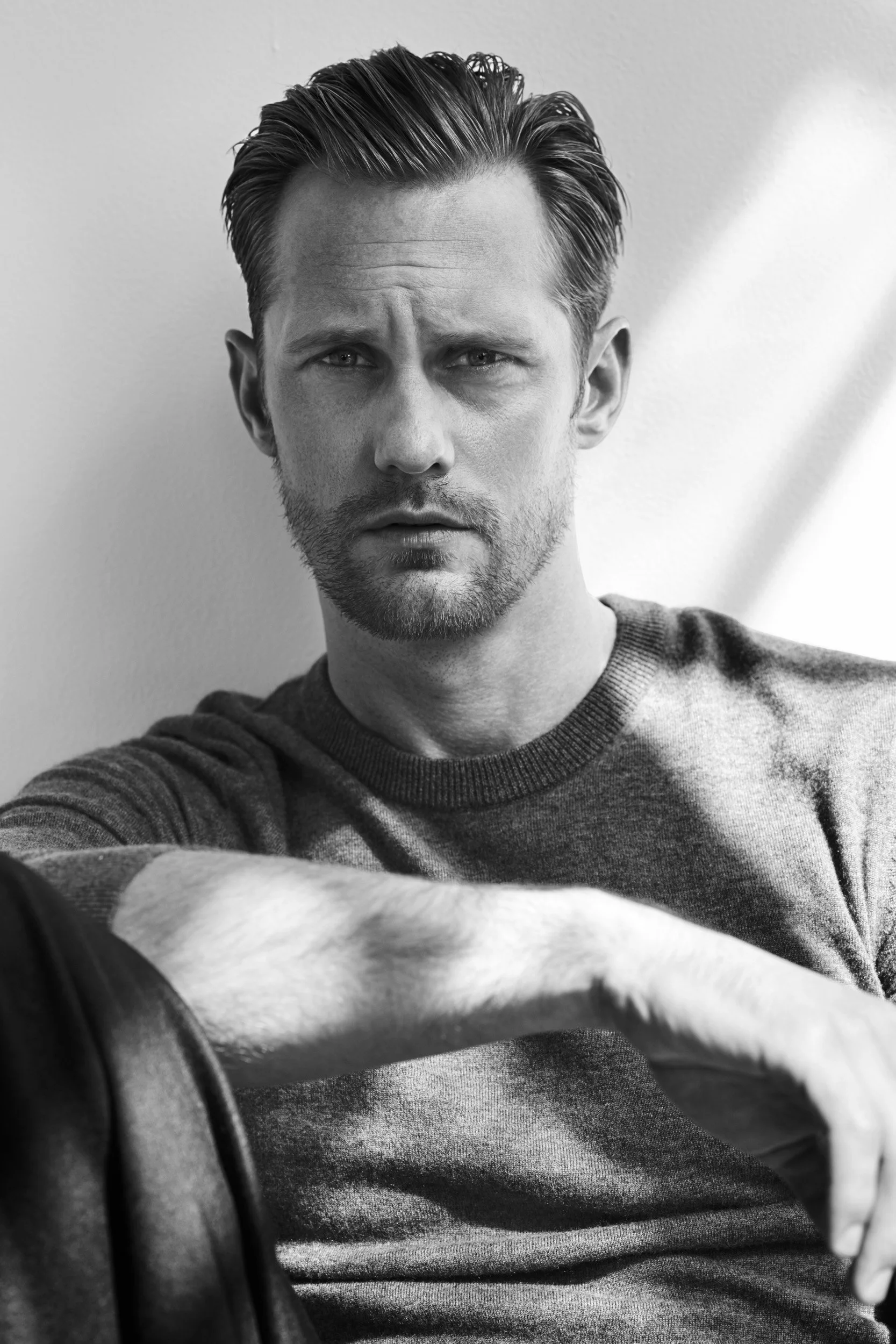 Black and white photo of a man with styled hair and a beard, looking serious, wearing a crew neck sweater, sitting against a plain wall.