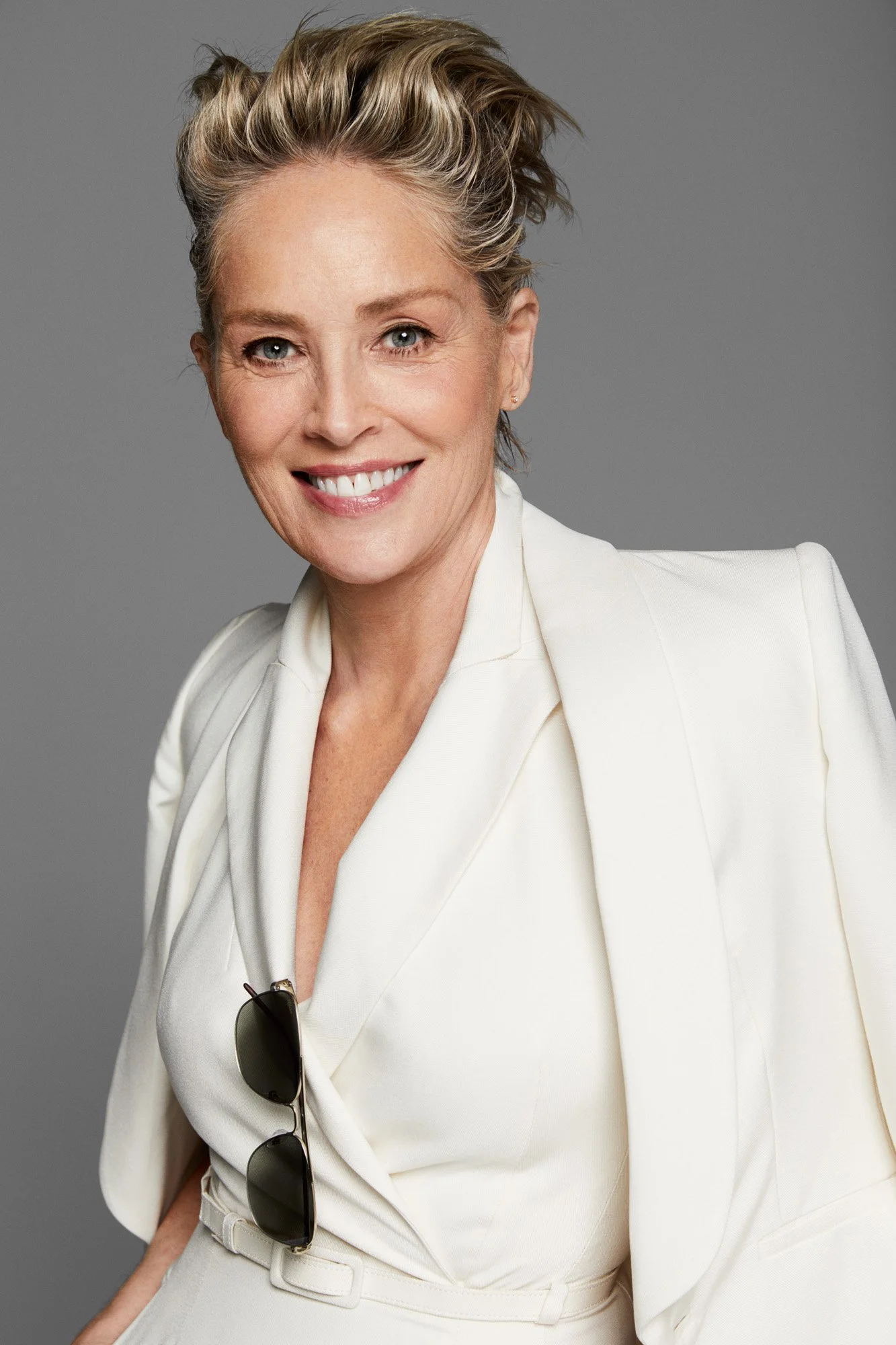 Smiling woman with short blonde hair wearing a white blazer and pants, with sunglasses hanging from the blazer, against a gray background.
