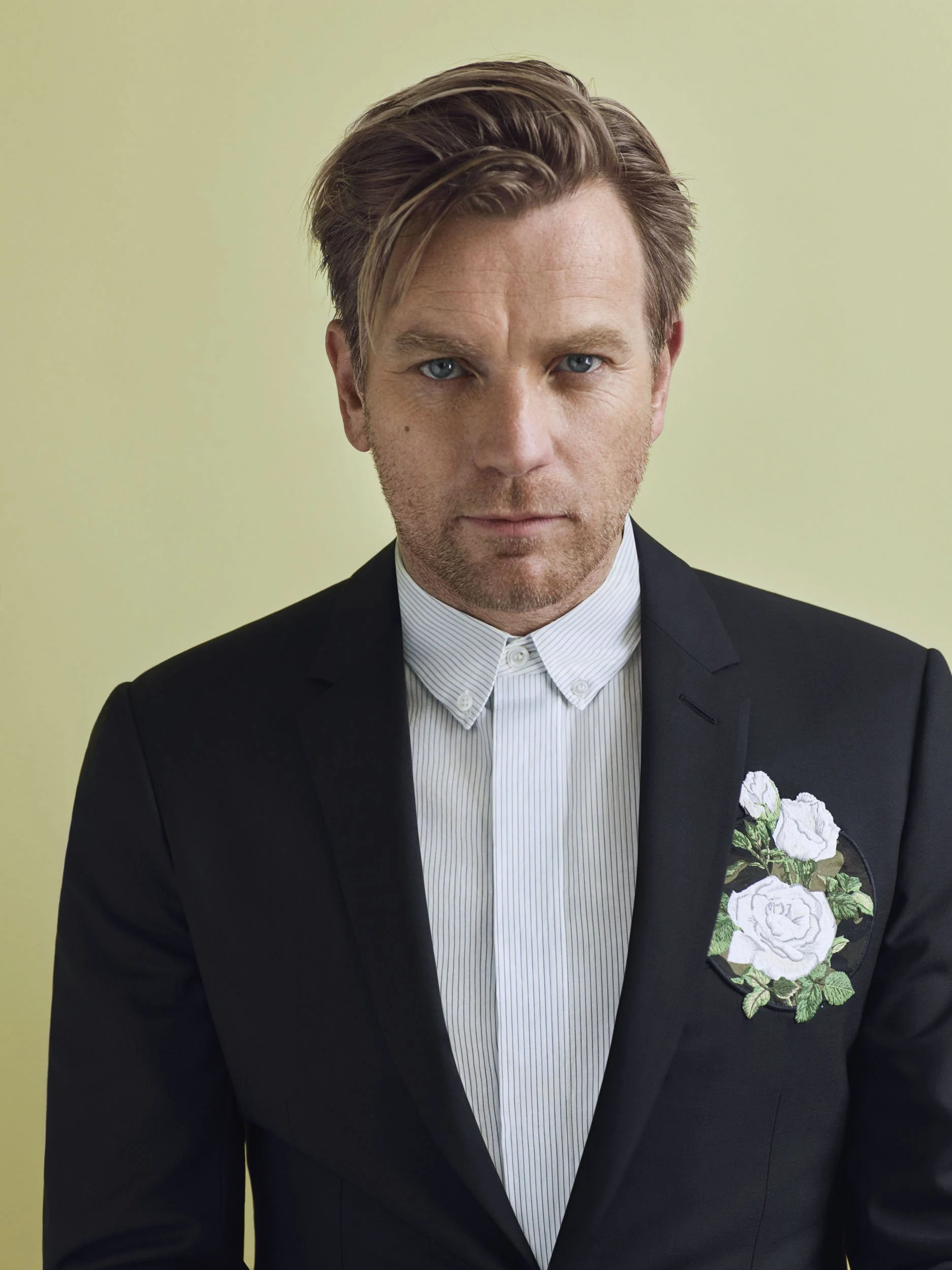 A man with light brown hair and blue eyes wearing a black suit jacket over a white dress shirt with a floral embroidery on the chest, standing against a plain light green background.