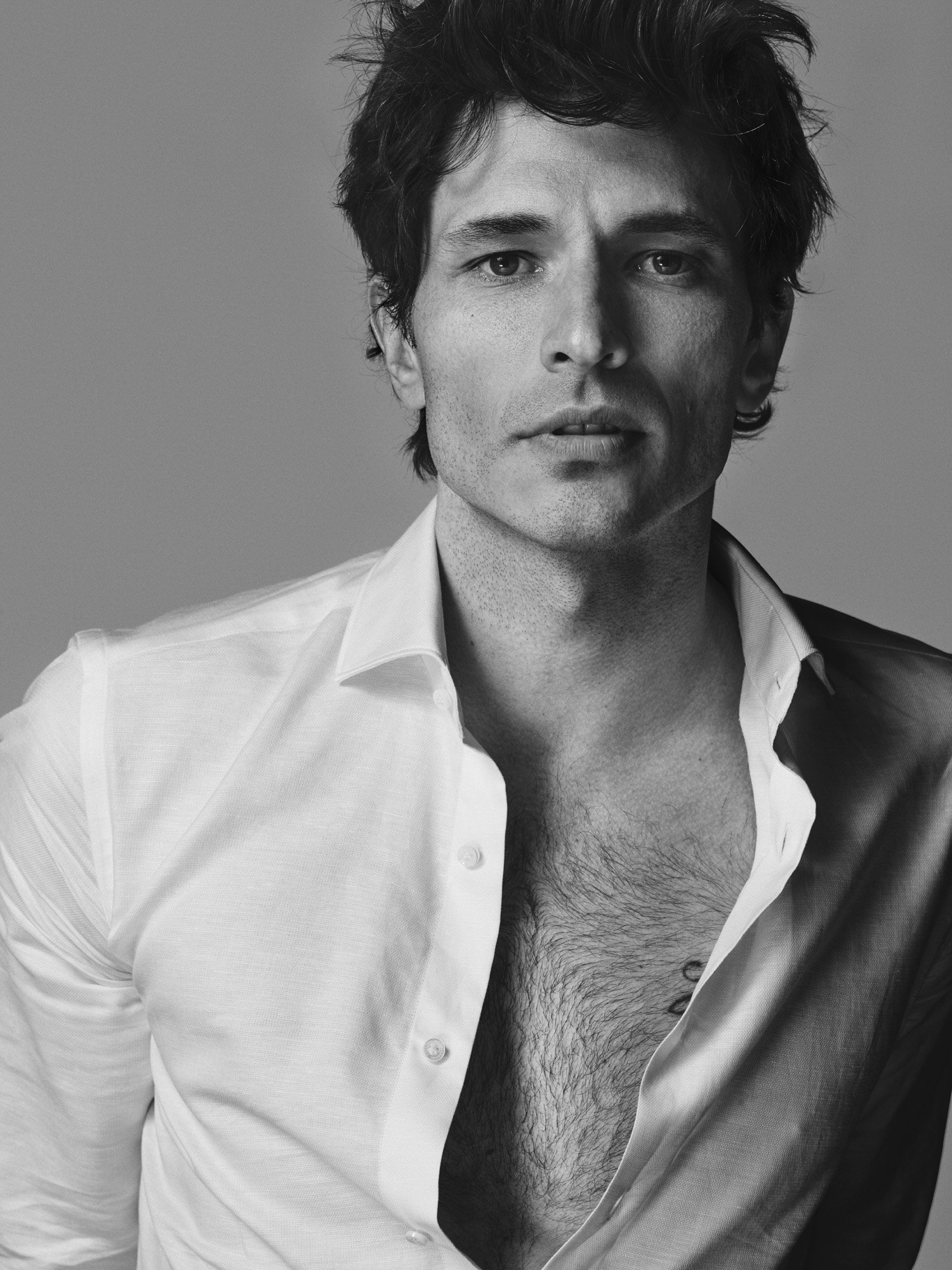 Black and white portrait of a man with dark, messy hair wearing an open white shirt, revealing chest hair, against a plain background.