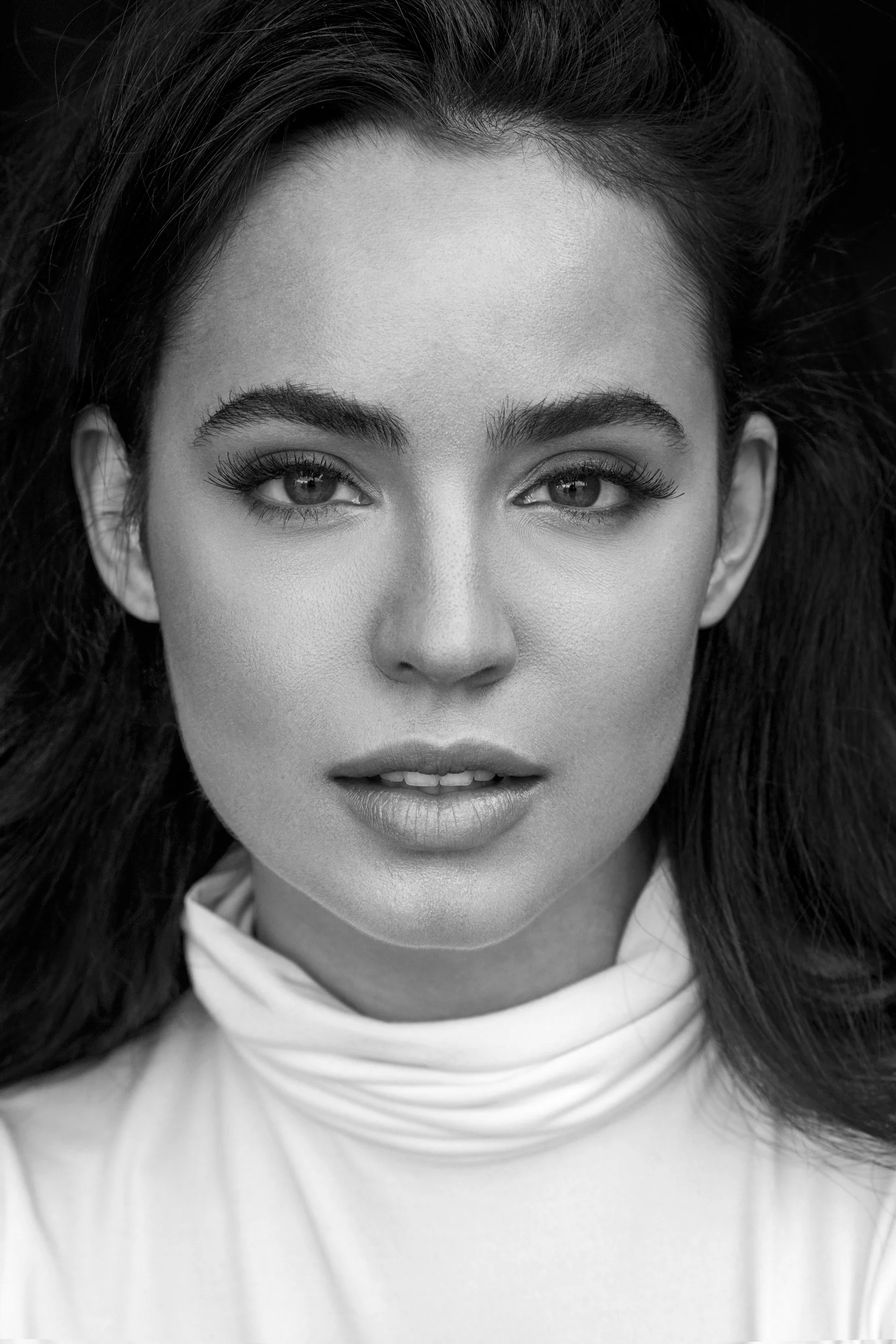 Close-up black and white photograph of a woman with long dark hair, high cheekbones, full lips, and light-colored eyes, wearing a high-necked white top.