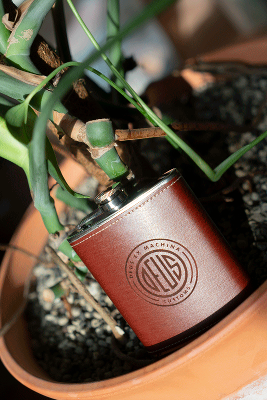 Flachmann mit Lederummantelung und lasergraviertem Deus Ex Machina Customs Logo in einem Pflanzentopf