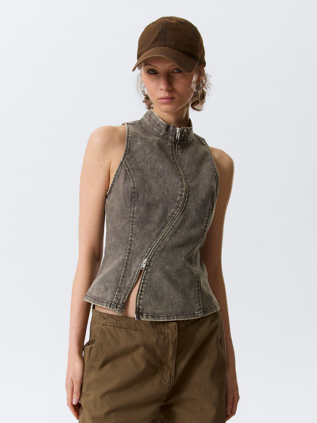 A woman wearing a brown cap, sleeveless zip-up gray denim vest, and khaki pants standing against a plain light gray background.