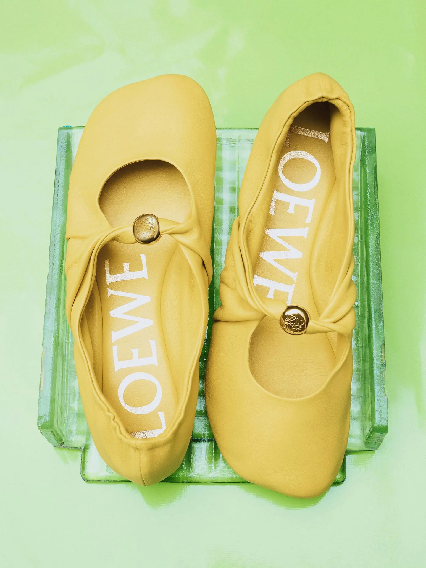 Pair of yellow Loewe shoes with bows and gold embellishments, placed on a green transparent display box.