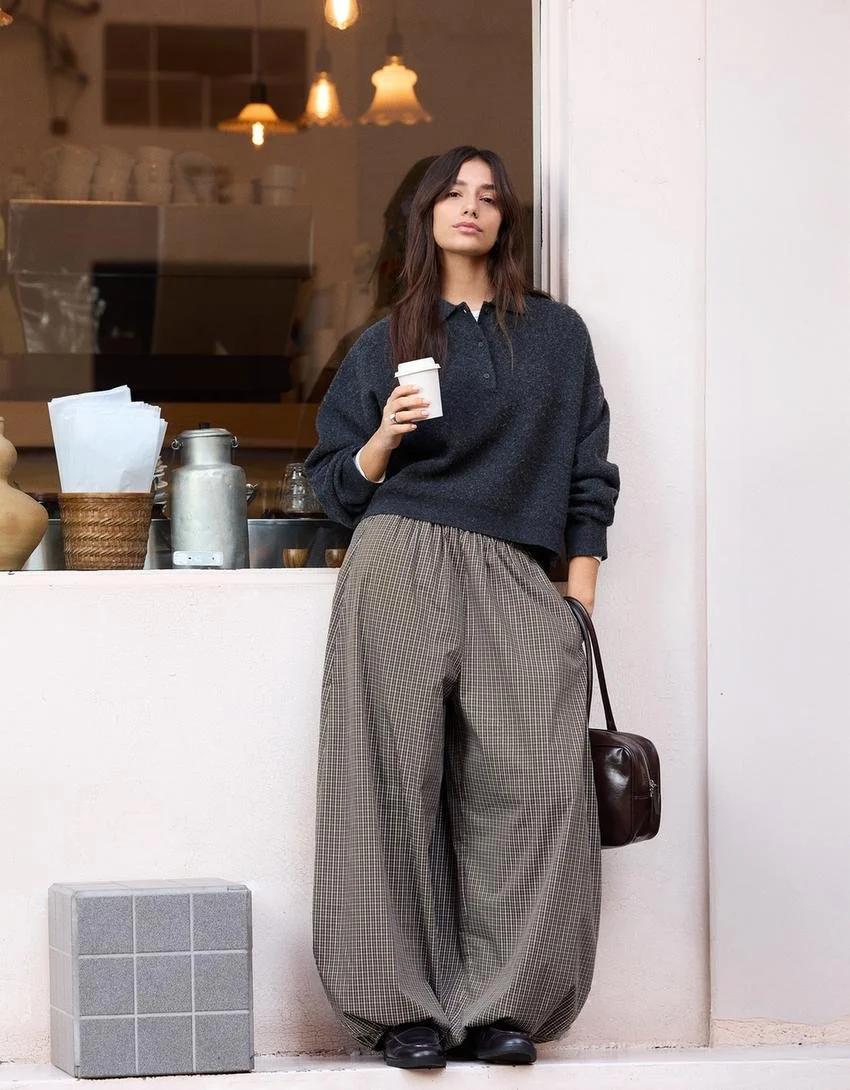 A young woman with shoulder-length brown hair and a neutral expression, wearing a dark sweater and loose checkered pants, holding a coffee cup in her right hand, standing outside a café with warm lighting inside, a black handbag hanging from her left arm, and a small square speaker on the floor.