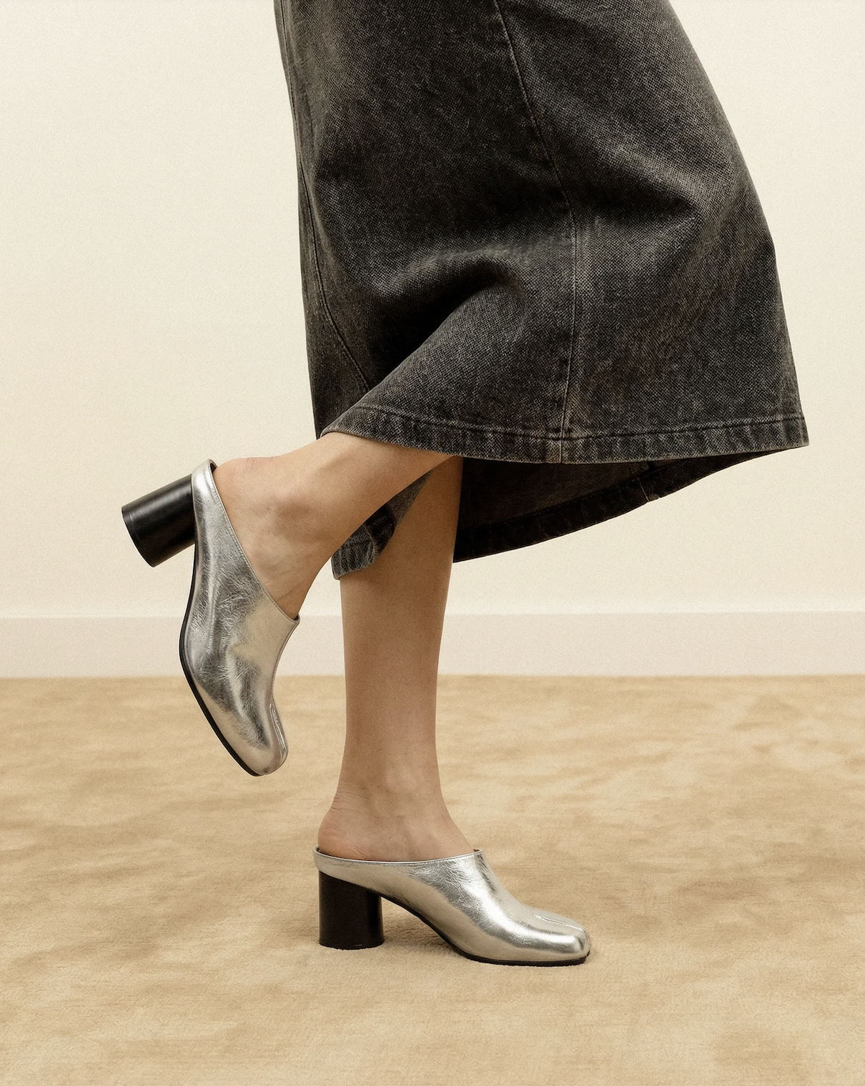 A person wearing metallic silver high-heeled shoes with black block heels and a black denim skirt, standing on a beige carpeted floor against a plain light-colored wall.