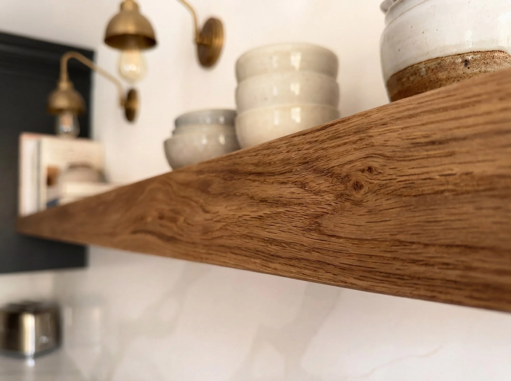 Close-up of a wood shelf with white ceramic bowls stacked on top, blurred background showing wall-mounted brass lamps and other kitchen items.