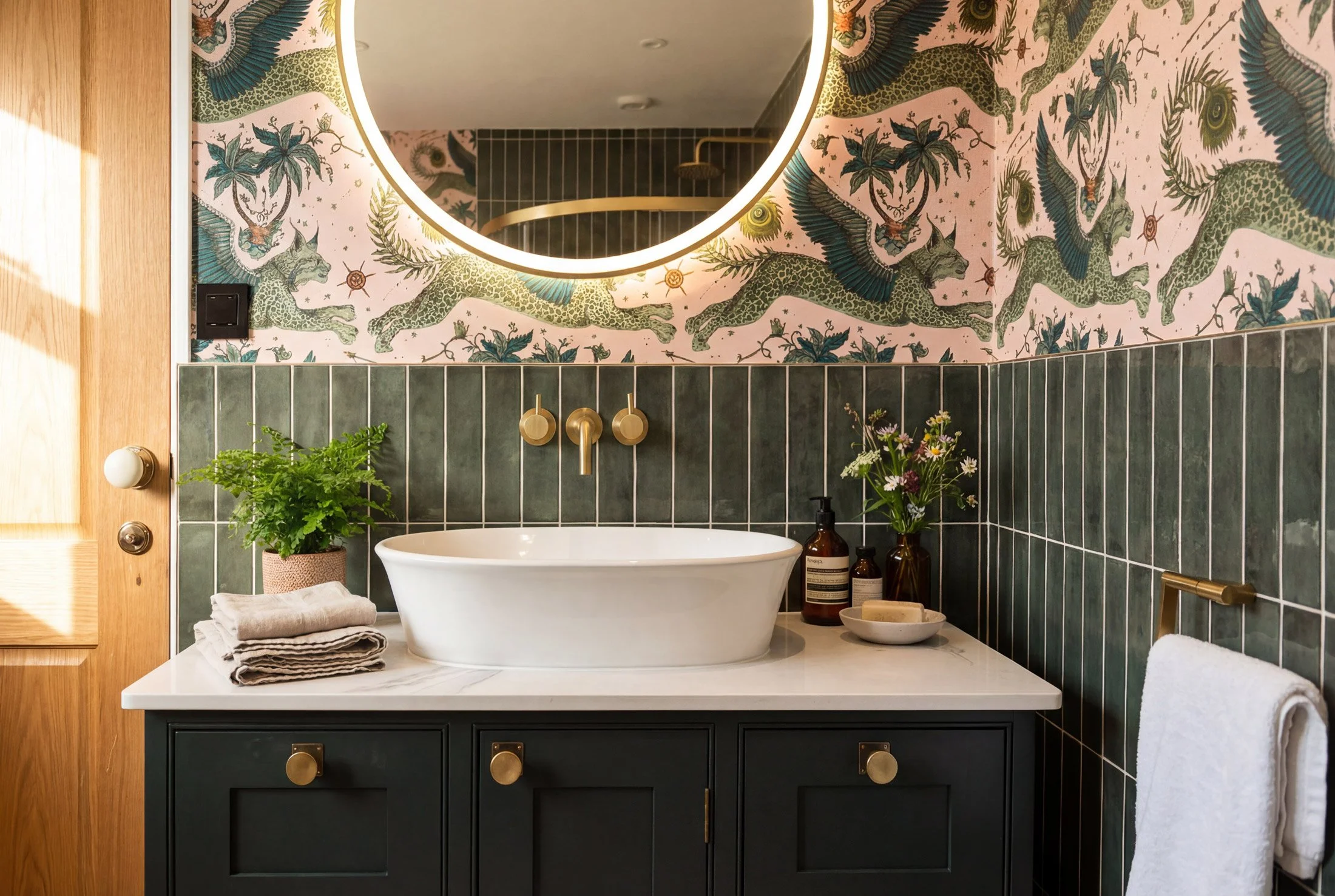 Modern bathroom with dark green tile walls, a white vessel sink, a round mirror with LED lighting, a black vanity with gold knobs, and decorative elements including a potted fern, folded towels, soap bottles, and a vase of flowers. A whimsical wallpaper with green lizards and plants decorates the upper wall.