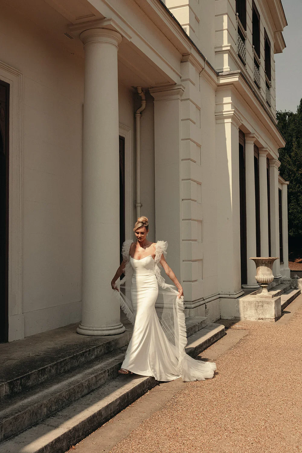 A woman in a white, elegant, form-fitting gown with ruffled shoulders standing on the steps of a large classical-style building with tall columns and architectural detailing.