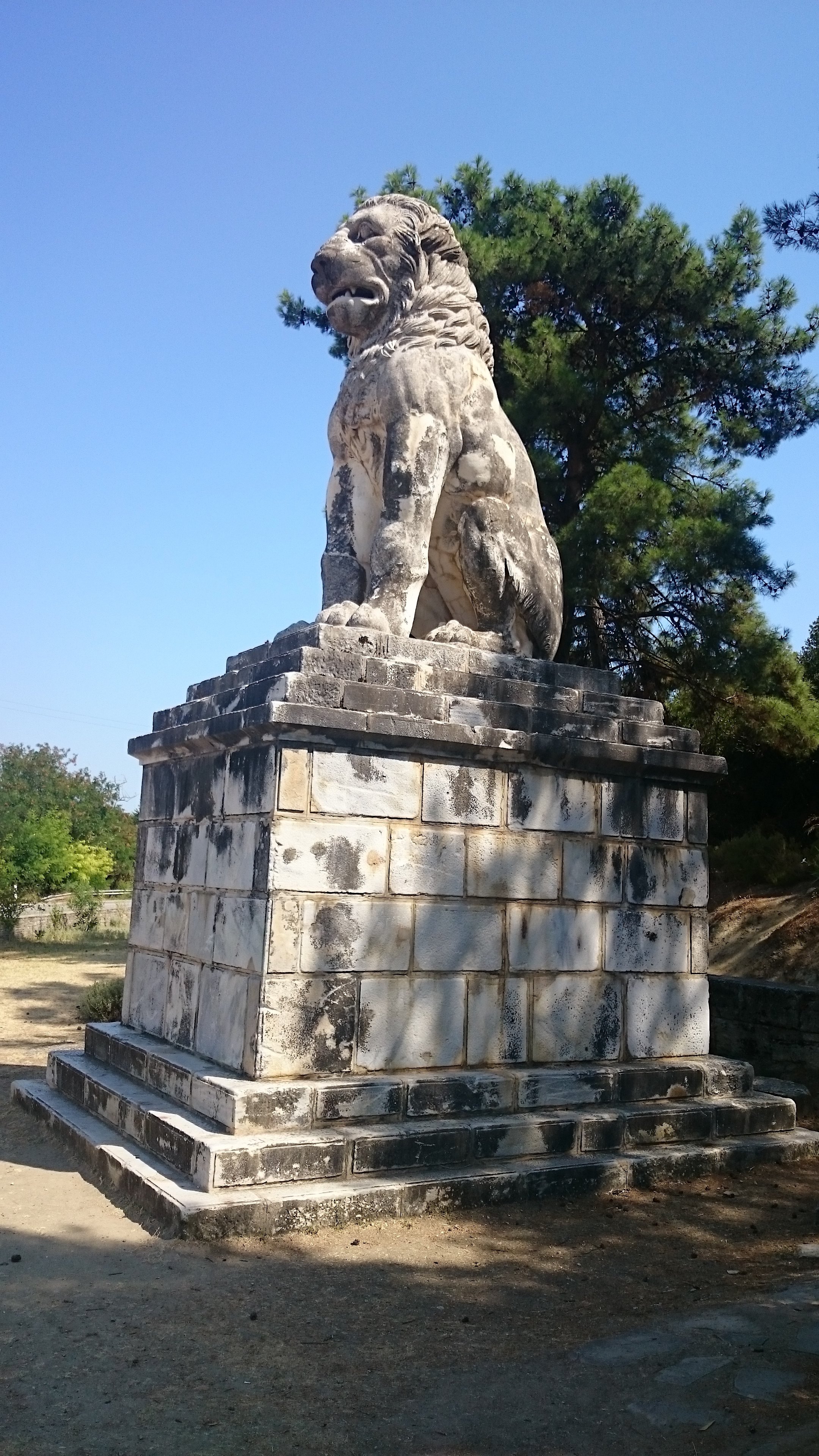 Ein steinerner Löwenbrunnen auf einem Podest, vor Bäumen und einem blauen Himmel.