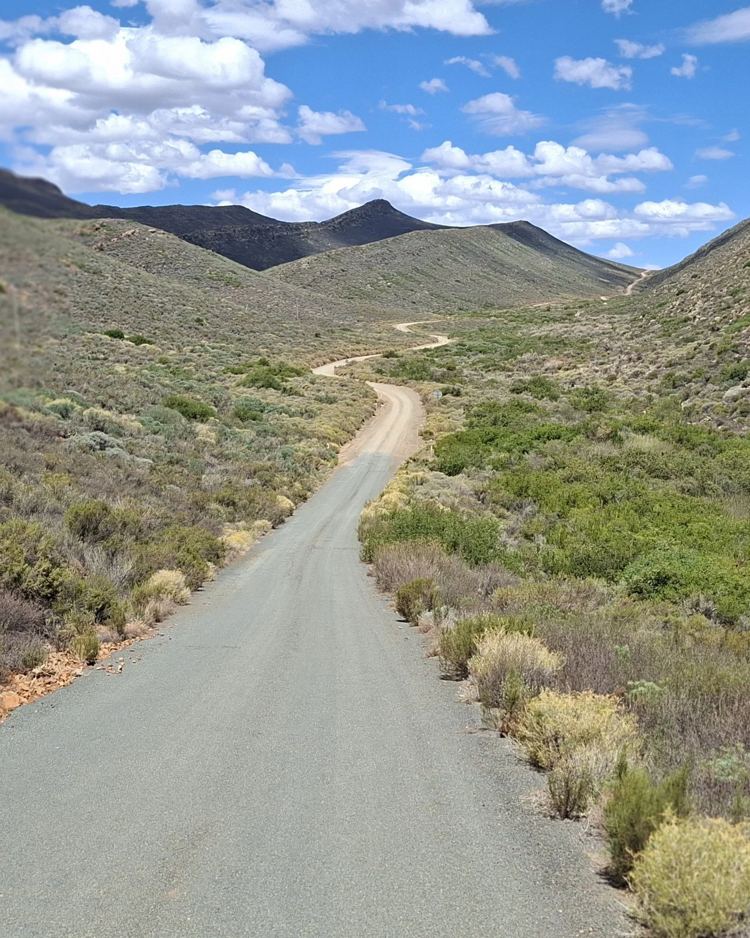 Route de terre sinueuse traversant un paysage désertique avec peu de végétation, entouré de collines et de montagnes sous un ciel partiellement nuageux.