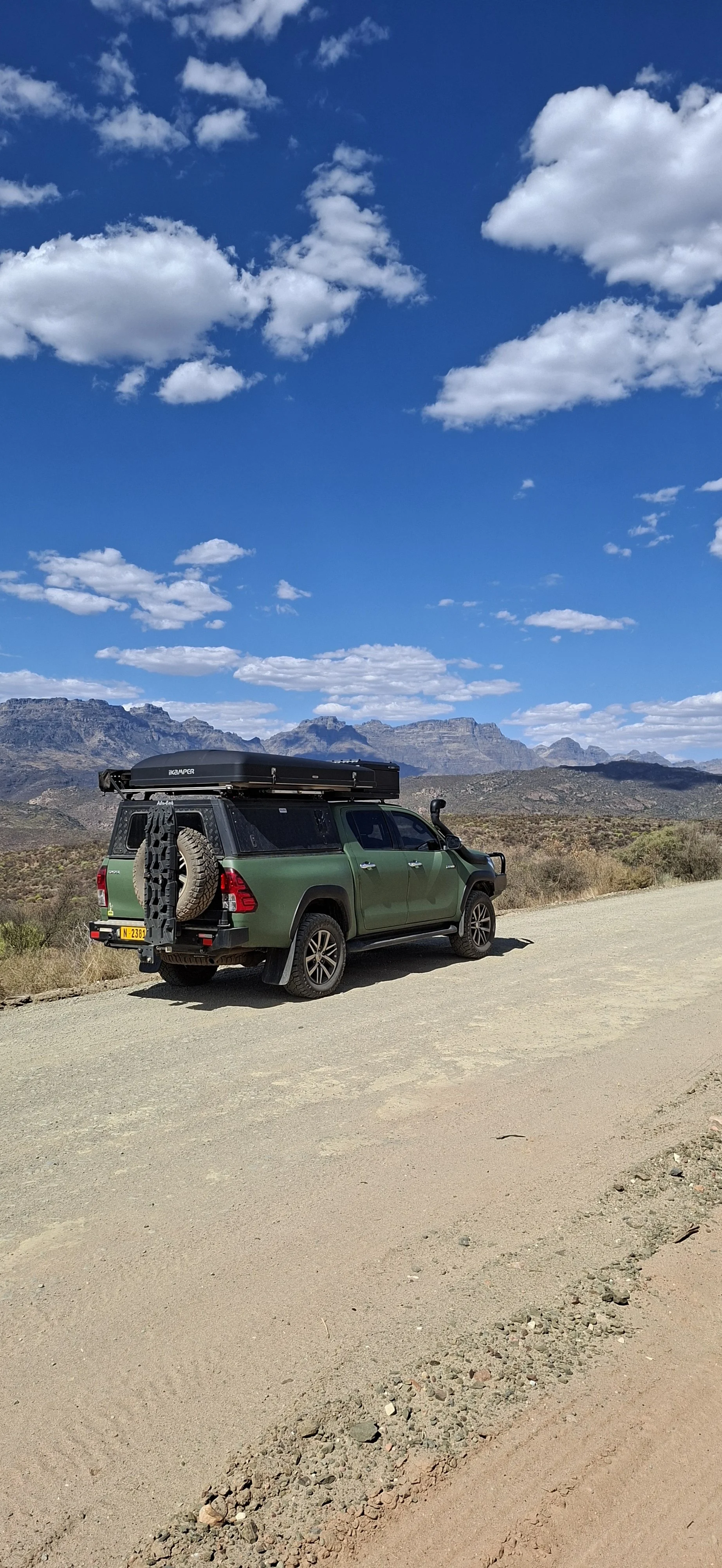 Voiture tout-terrain verte avec un coffre de toit et une roue de secours attachée, garée sur une route désertique dans un paysage montagneux, ciel bleu avec des nuages blancs.