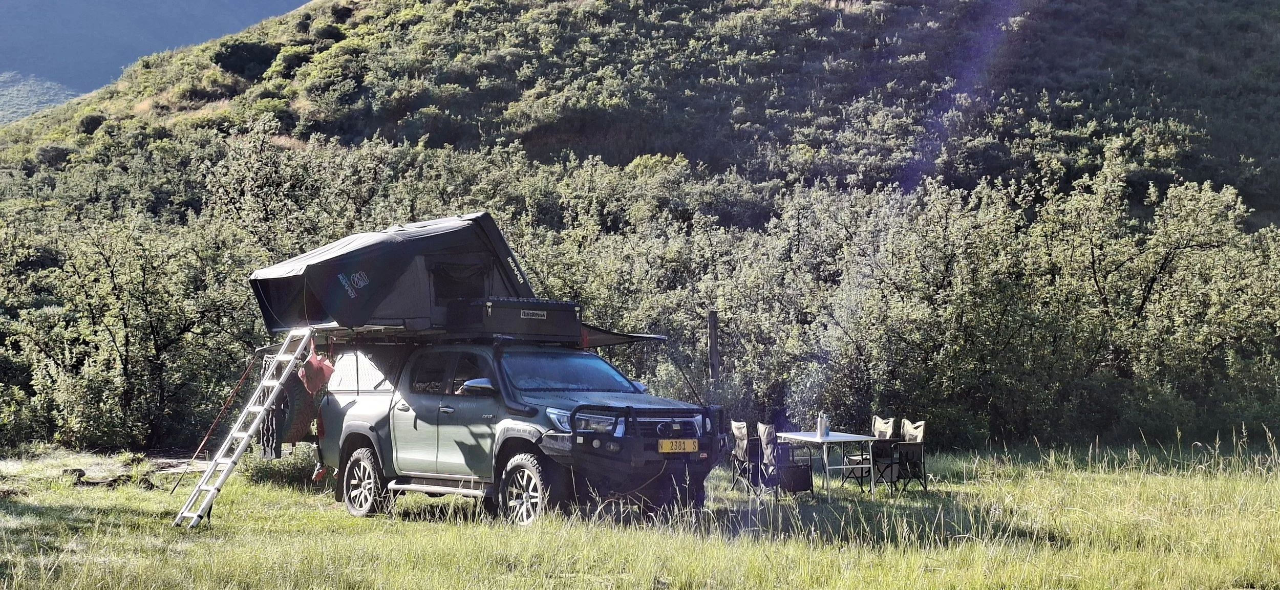 Un véhicule de camping avec un toit relevable dans une zone boisée et ensoleillée, avec une table et des chaises à l'extérieur.