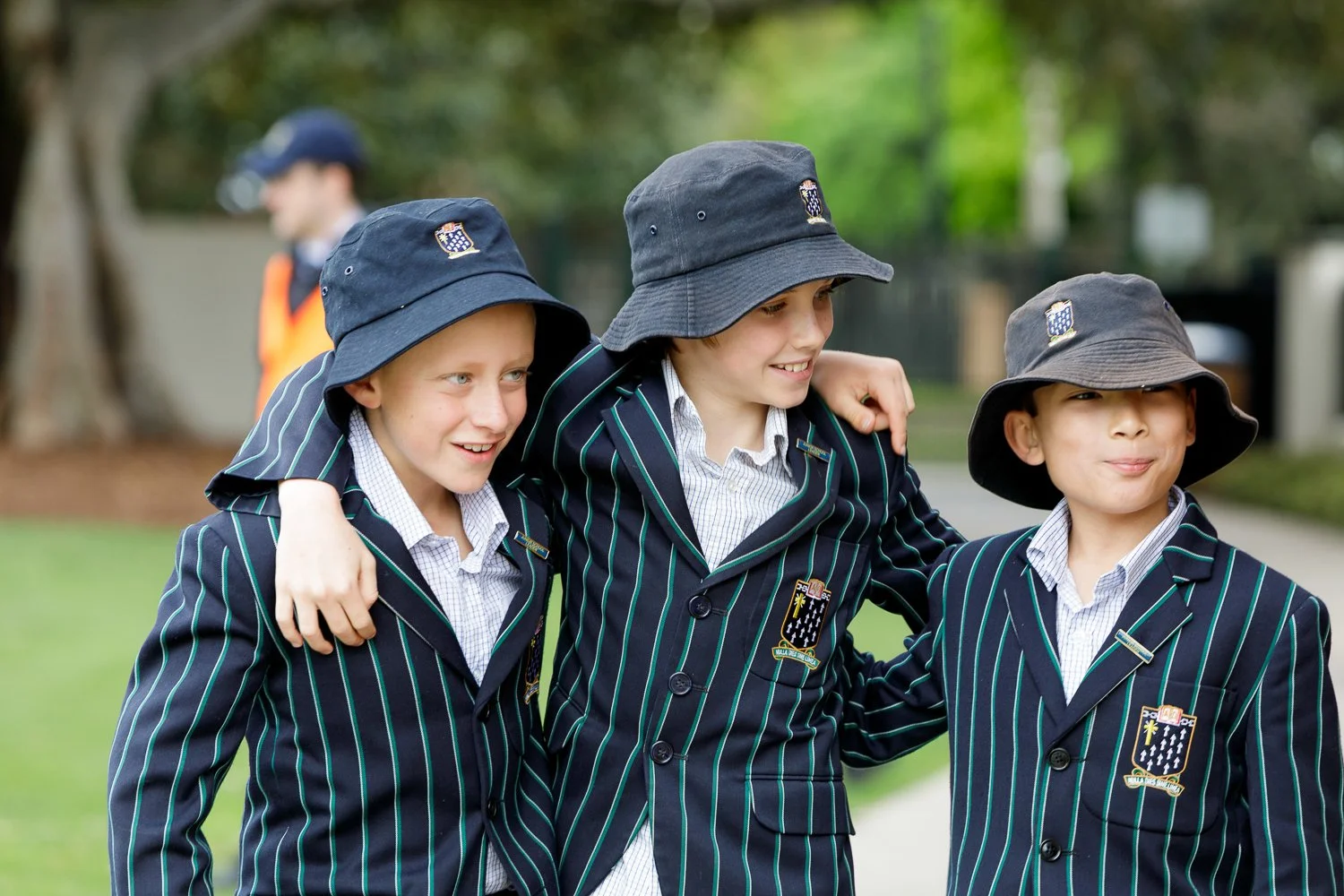 Three young boys in school uniforms with striped blazers and bucket hats, smiling and walking together with their arms around each other's shoulders in a park or outdoor area.