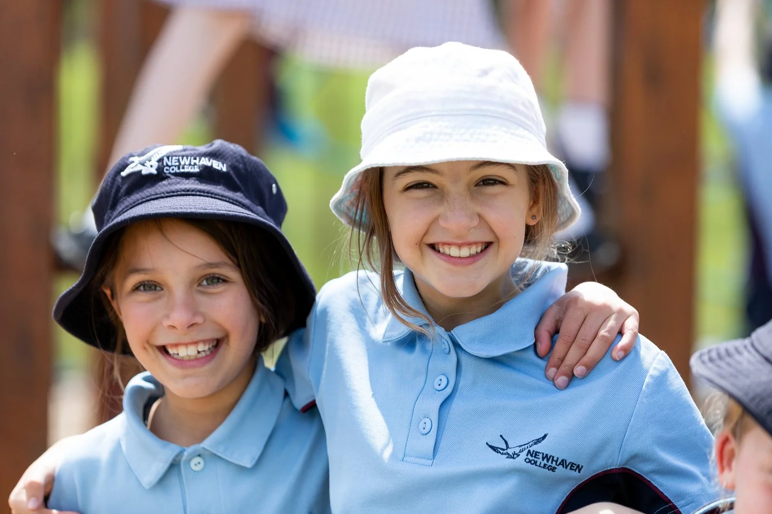 Two smiling young girls in blue shirts with 'Newhaven College' logos, wearing hats, standing outdoors, with a wooden structure in the background.
