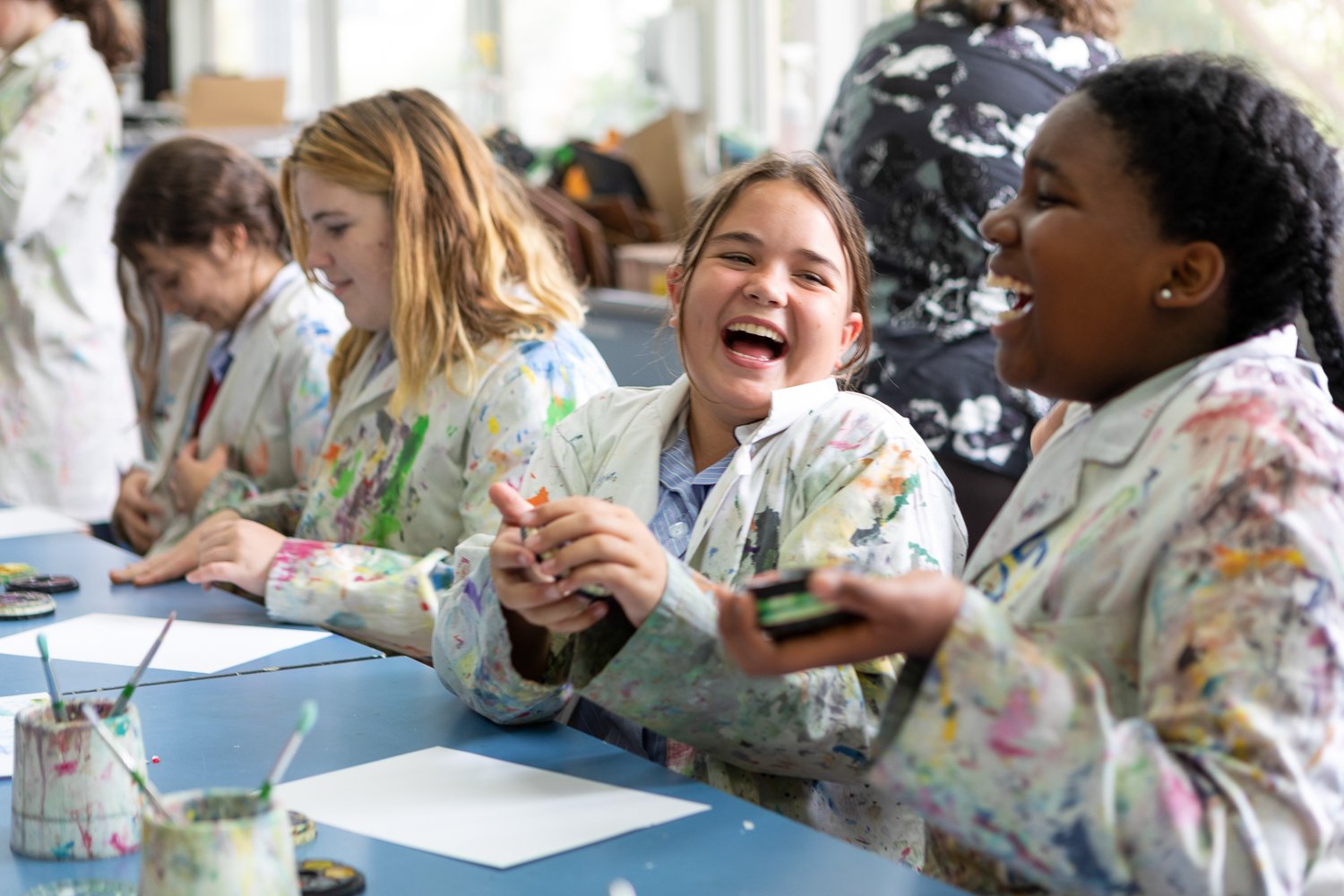 Group of young girls in paint-splattered smocks sitting at a table, laughing and interacting in an art classroom. school photography and advertising photography, content creation and video by Jess Grant and Alys Carr, Melbourne Australia.