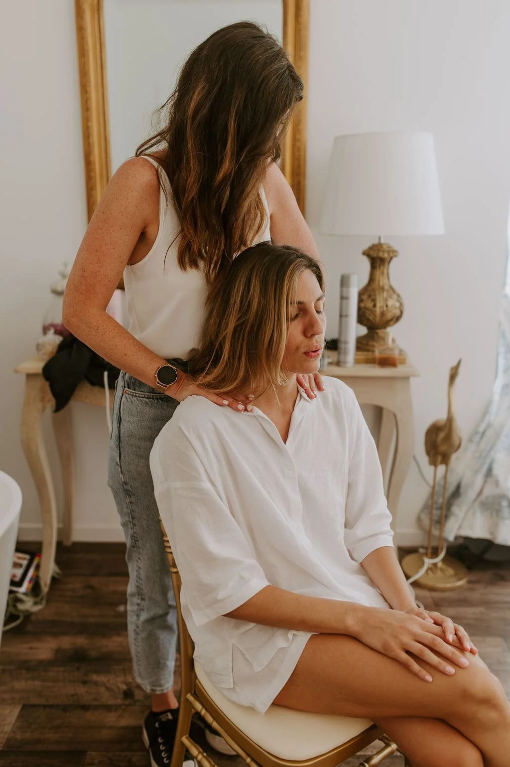 Deux femmes dans un intérieur chaleureux. Une femme aux cheveux longs bruns, assise, en chemise blanche, reçoit une massage ou un soin du cou de la part d'une autre femme aux cheveux longs bruns, qui se tient derrière elle.
