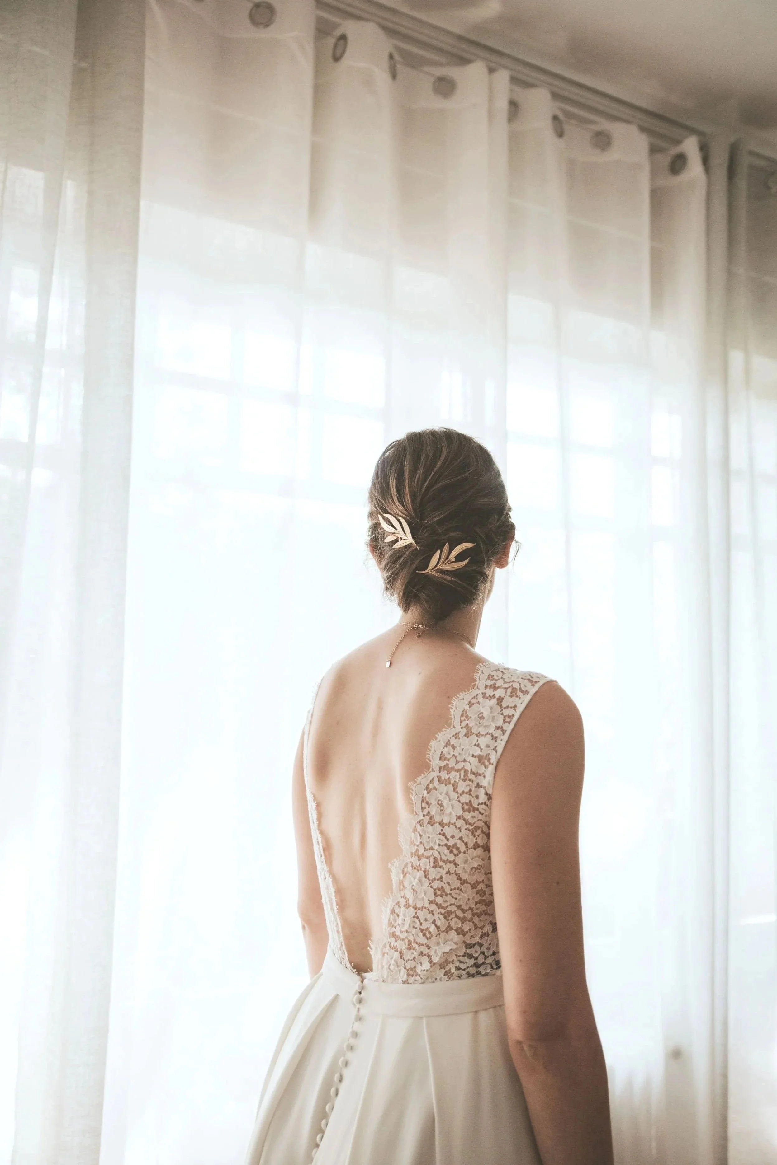 Une femme vue de dos portant une robe de mariée en dentelle, les cheveux coiffés en chignon avec des accessoires ornés de feuilles, devant une fenêtre avec des rideaux blancs en voile.