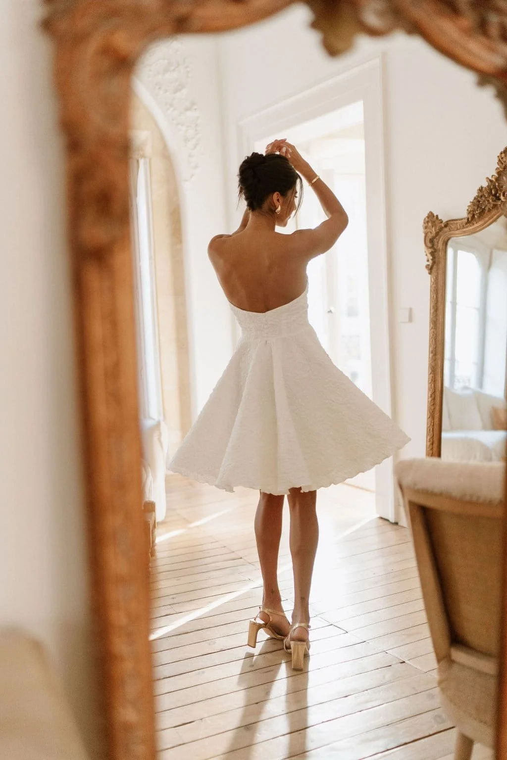 Une femme regarde dans un miroir en portant une robe blanche courte et des talons hauts, dans une pièce lumineuse.