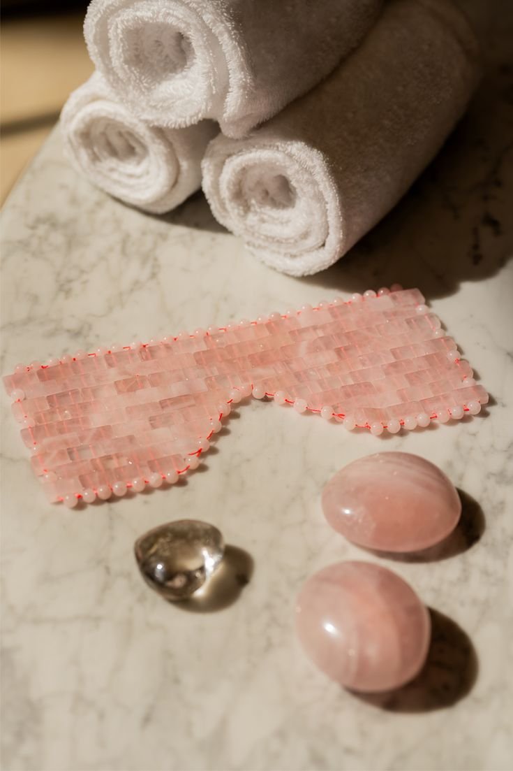 Trois serviettes blanches roulées, deux pierres roses, une pierre argentée, une bande de perles roses sur une surface en marbre.