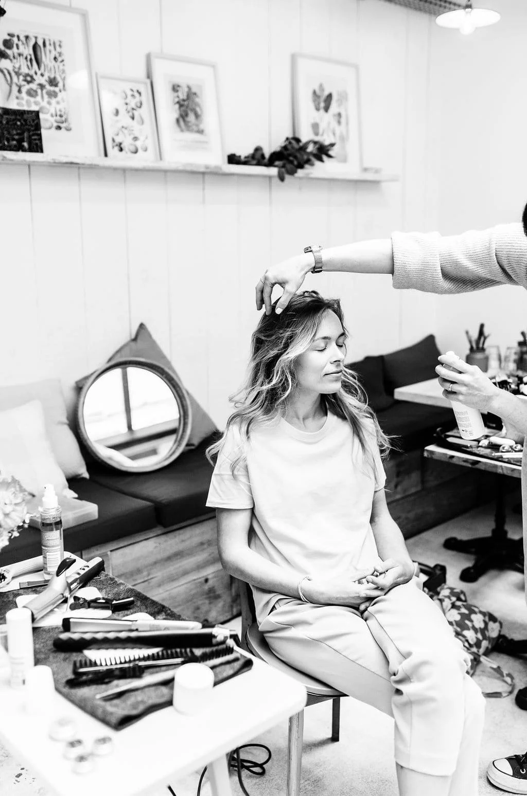 Une femme est assise en train de recevoir un soin capillaire ou un maquillage dans un salon. Un professionnel utilise un spray ou un produit sur ses cheveux ou son visage, dans un environnement décoré de tableaux et d'accessoires de beauté.
