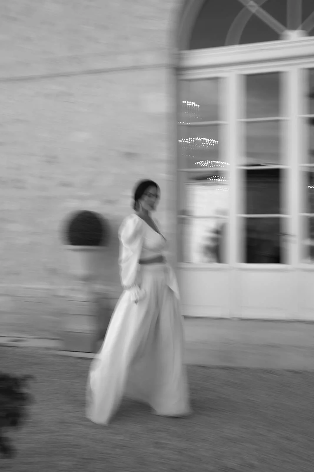 Une femme en robe longue et vêtement clair, debout près d'un bâtiment avec une façade en pierre et des fenêtres, dans un contexte urbain, en noir et blanc avec un effet de flou artistique.