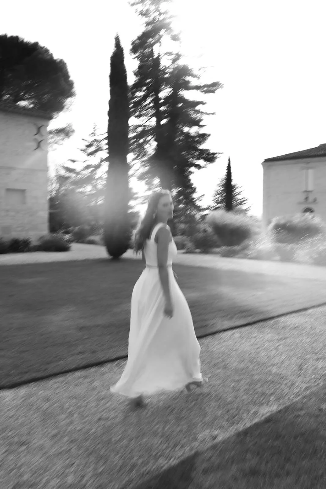 Une femme en robe blanche marche dans une rue résidentielle, avec des arbres et des maisons en arrière-plan, en lumière de fin d'après-midi.