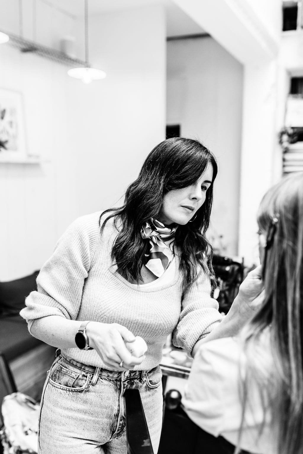 Une femme aux cheveux longs et foncés, portant un pull clair, une écharpe et une montre, est en train de maquiller ou d'appliquer un soin à une autre personne dans un espace intérieur.