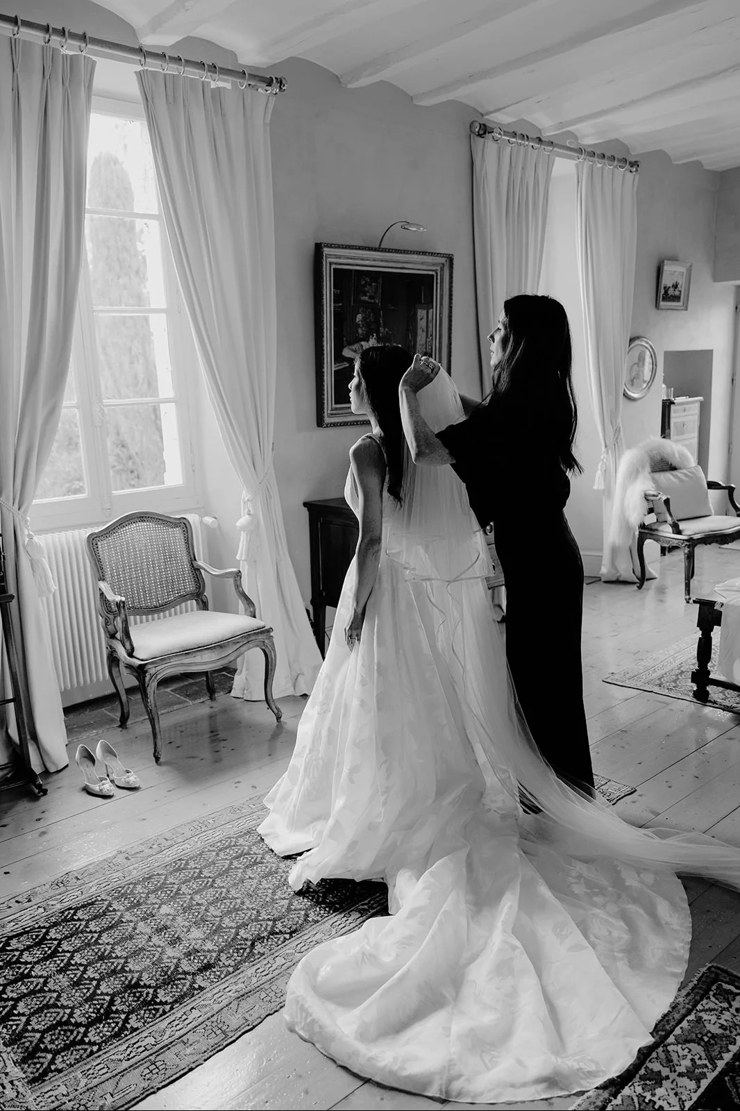 Femme habillée en mariée avec une robe blanche, assistée par une autre femme selon la préparation dans une pièce lumineuse, avec des rideaux blancs, des meubles anciens et des chaussures à talons à côté d'une chaise.